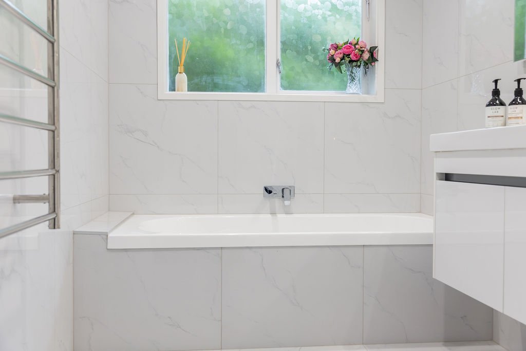Bathroom with a white bathtub, a window with green foliage outside, a potted pink flower arrangement, and soap dispensers on a white sink counter.