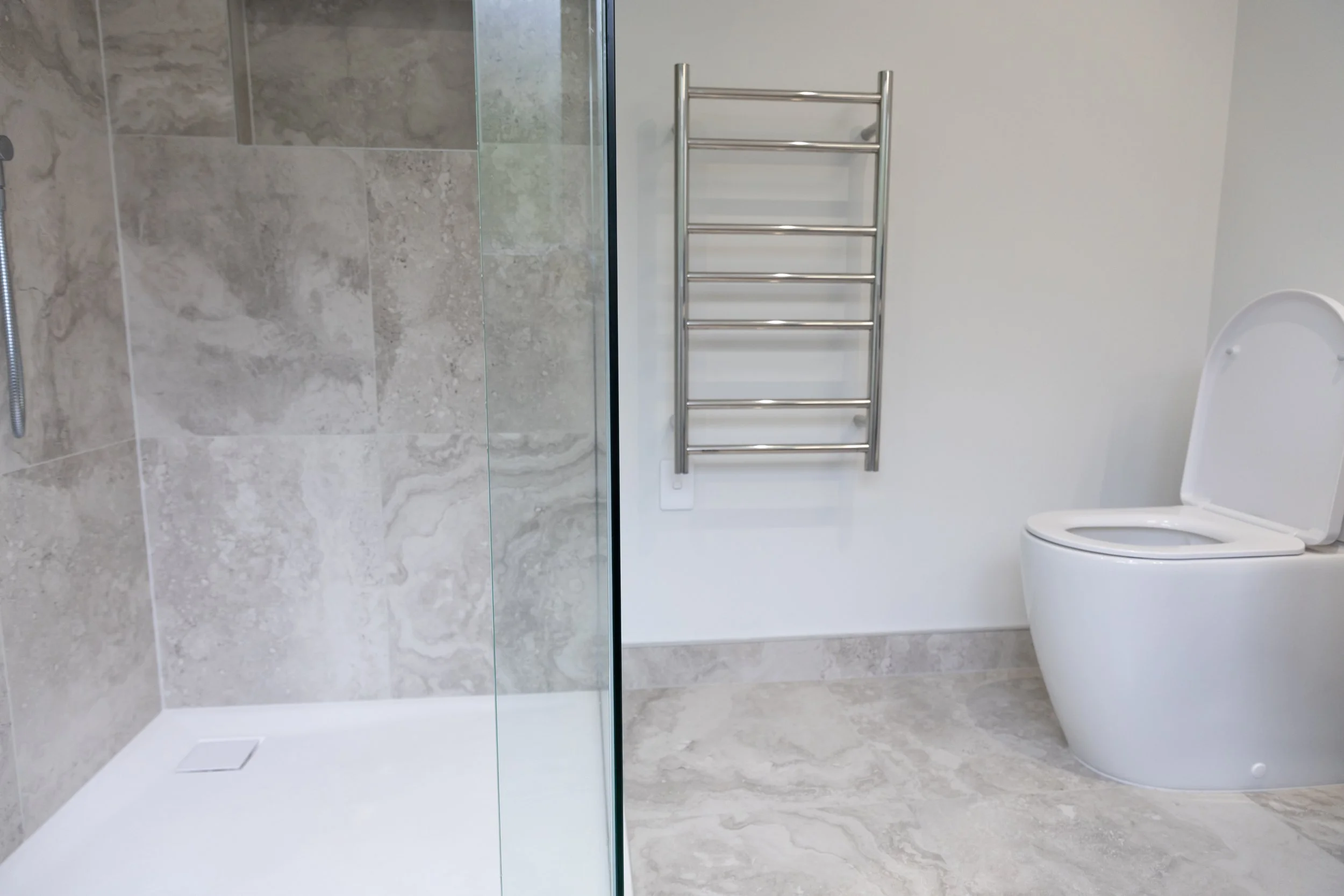 Modern bathroom with a glass-enclosed shower, beige marble floors, a heated towel rack, and a white toilet with the lid open.