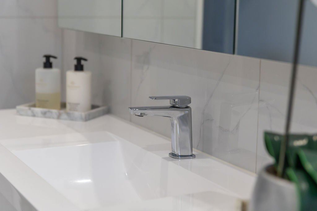 A modern bathroom sink with a chrome faucet, a marble tray with soap dispensers, and a plant partially visible on the right side.