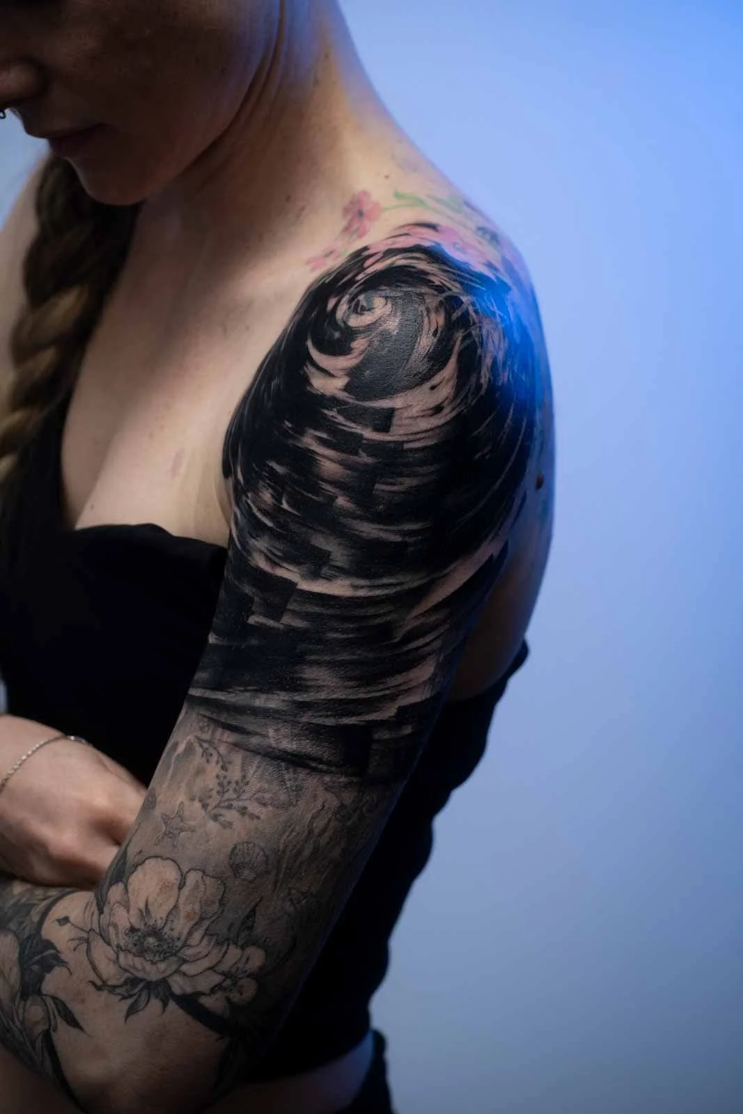 Close-up of a woman's shoulder and upper arm with a black spiral tattoo, part of a floral tattoo, and some pink flowers near her neck. She has light skin and braided hair.