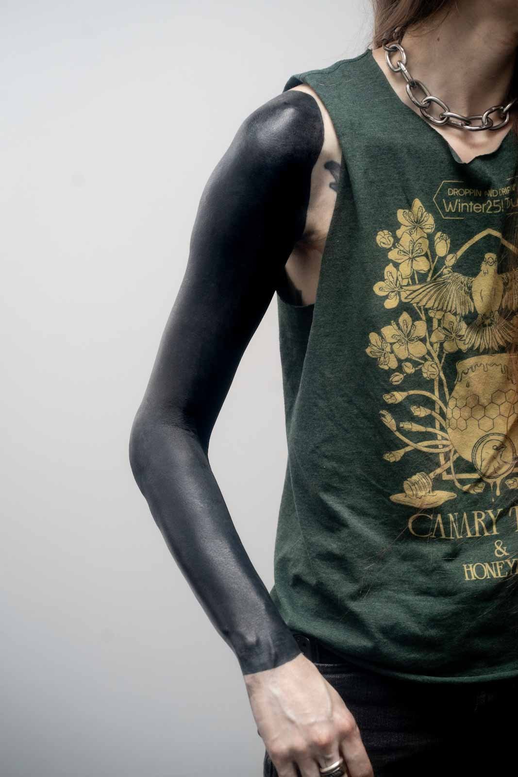 Person wearing a sleeveless T-shirt with graphic and text, wearing a chain necklace, and having a blackout sleeve tattoo on their left arm.
