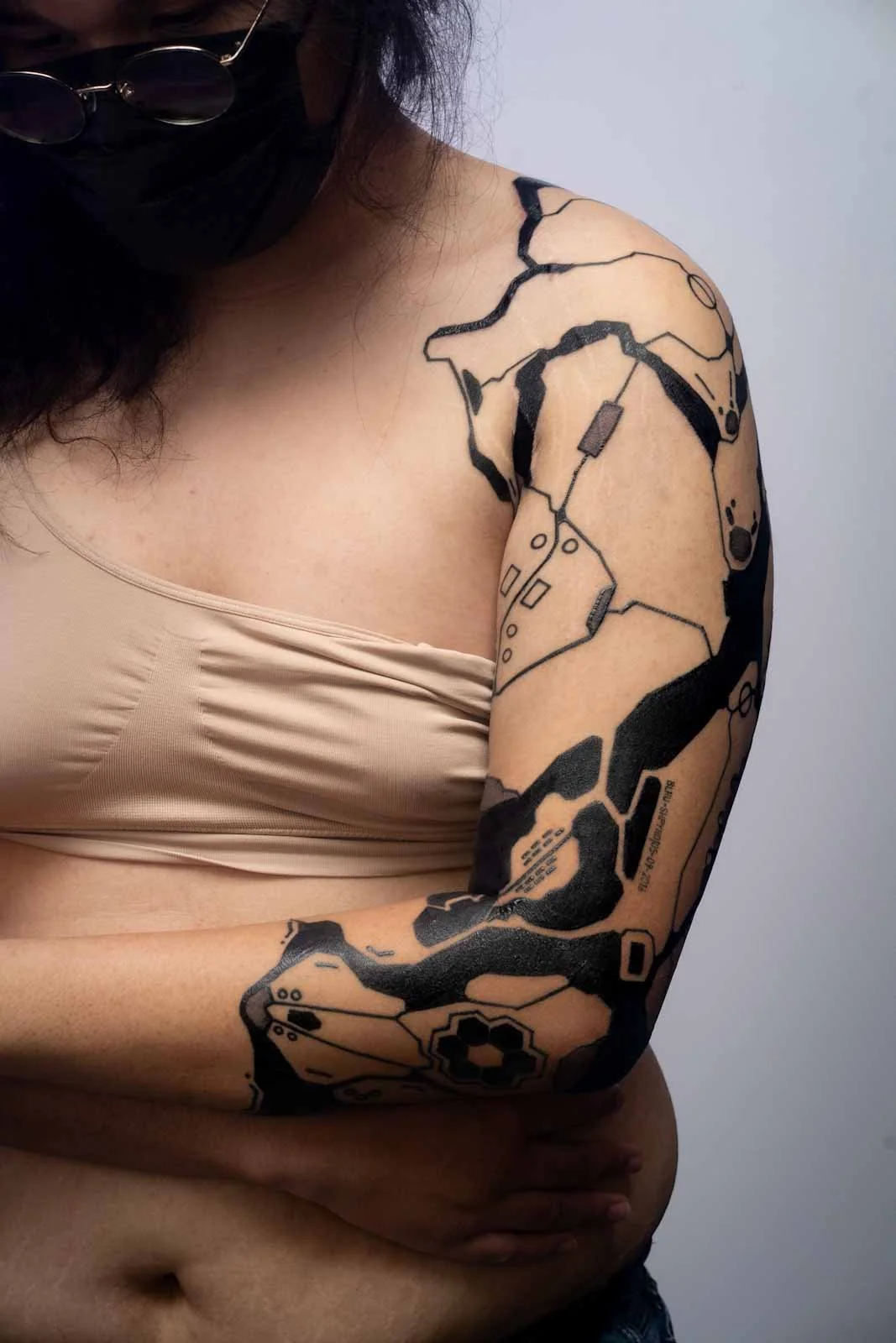 Close-up of a person's shoulder and upper arm with a black tattoo resembling a cybernetic prosthesis, wearing sunglasses and a beige top.