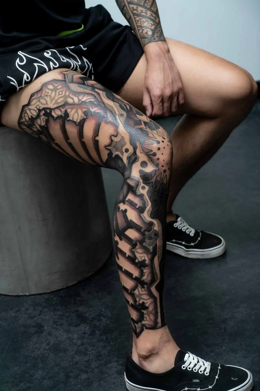 A person with a large, detailed black and gray tattoo sleeve on their right leg, sitting on a gray piece of furniture, wearing black Vans sneakers, black shorts, and a black shirt.