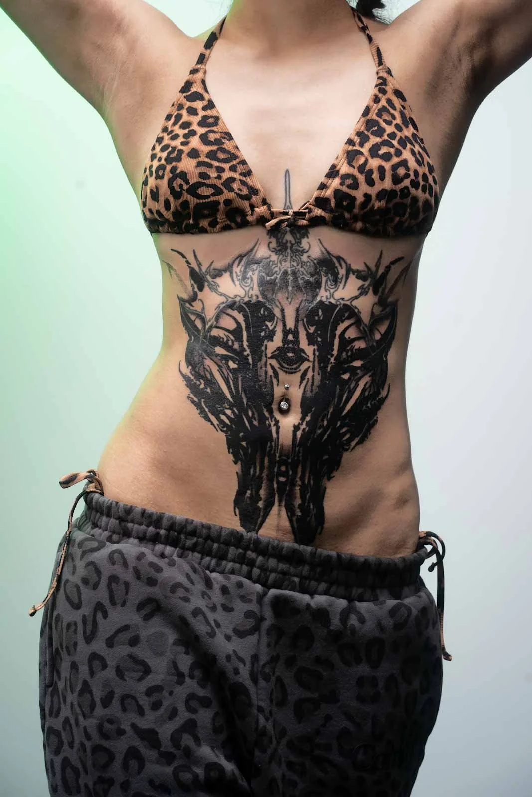 Person wearing a leopard print bikini top and loose animal print shorts, revealing an intricate large black tribal-style tattoo of an abstract face on the stomach, with belly button jewelry. Arms raised above head.