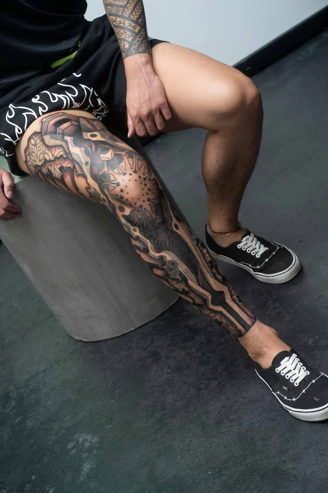 A person is sitting on a gray, round stool with one leg bent and the other extended. They are wearing black sneakers  and have a detailed, large tattoo covering theirleg, featuring abstract and geometric designs. They are dressed in a black t-shirt a