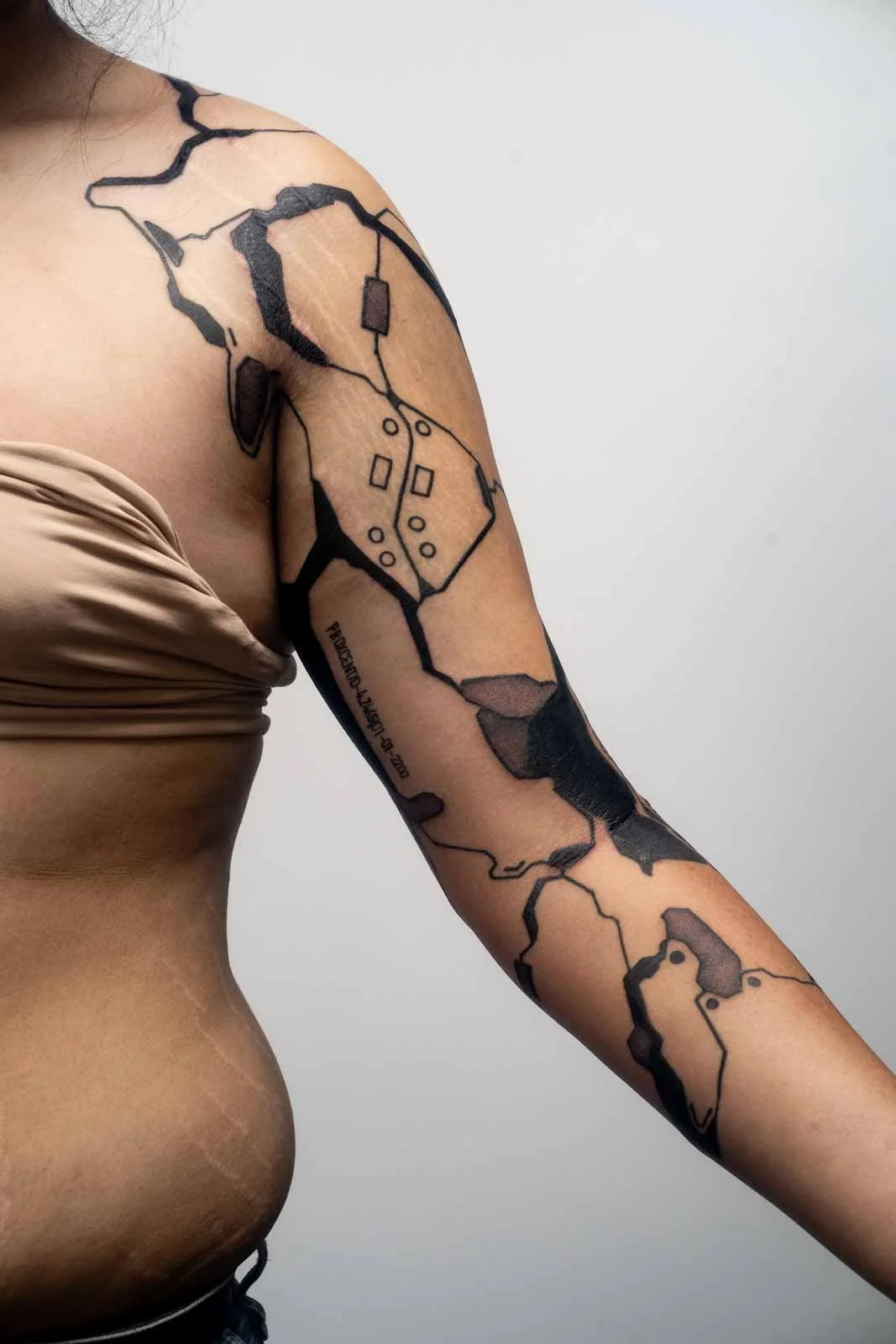 Tattoo of a cyberware prosthesis on the arm of someone wearing a being top on a white background. 