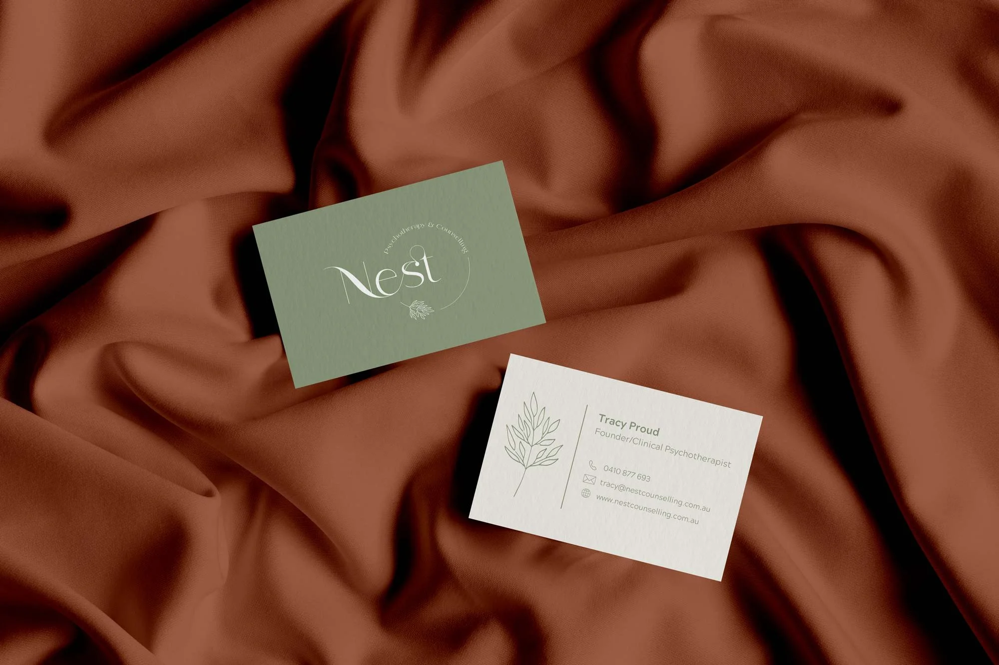 Two business cards on a brown satin fabric background. One card is green with white text, reading 'Nest' and 'Psychotherapy & Counselling.' The other card is white with green text, displaying contact information for Tracy Proud, a founder and clinical psychotherapist, with a simple line drawing of a plant.