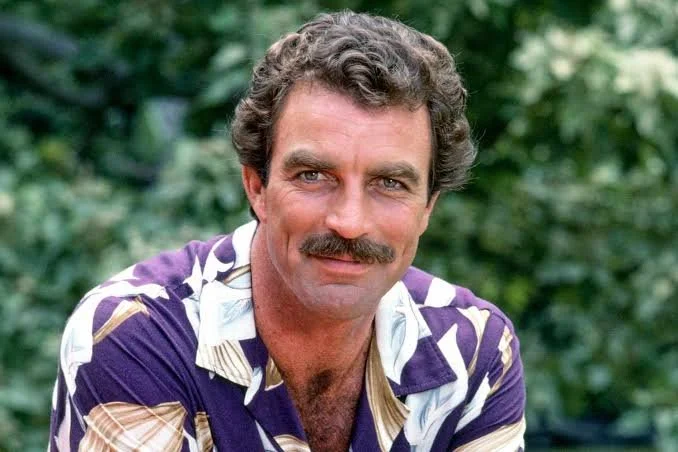 Man with curly hair and a mustache wearing a colorful Hawaiian shirt, outdoors with green foliage background.