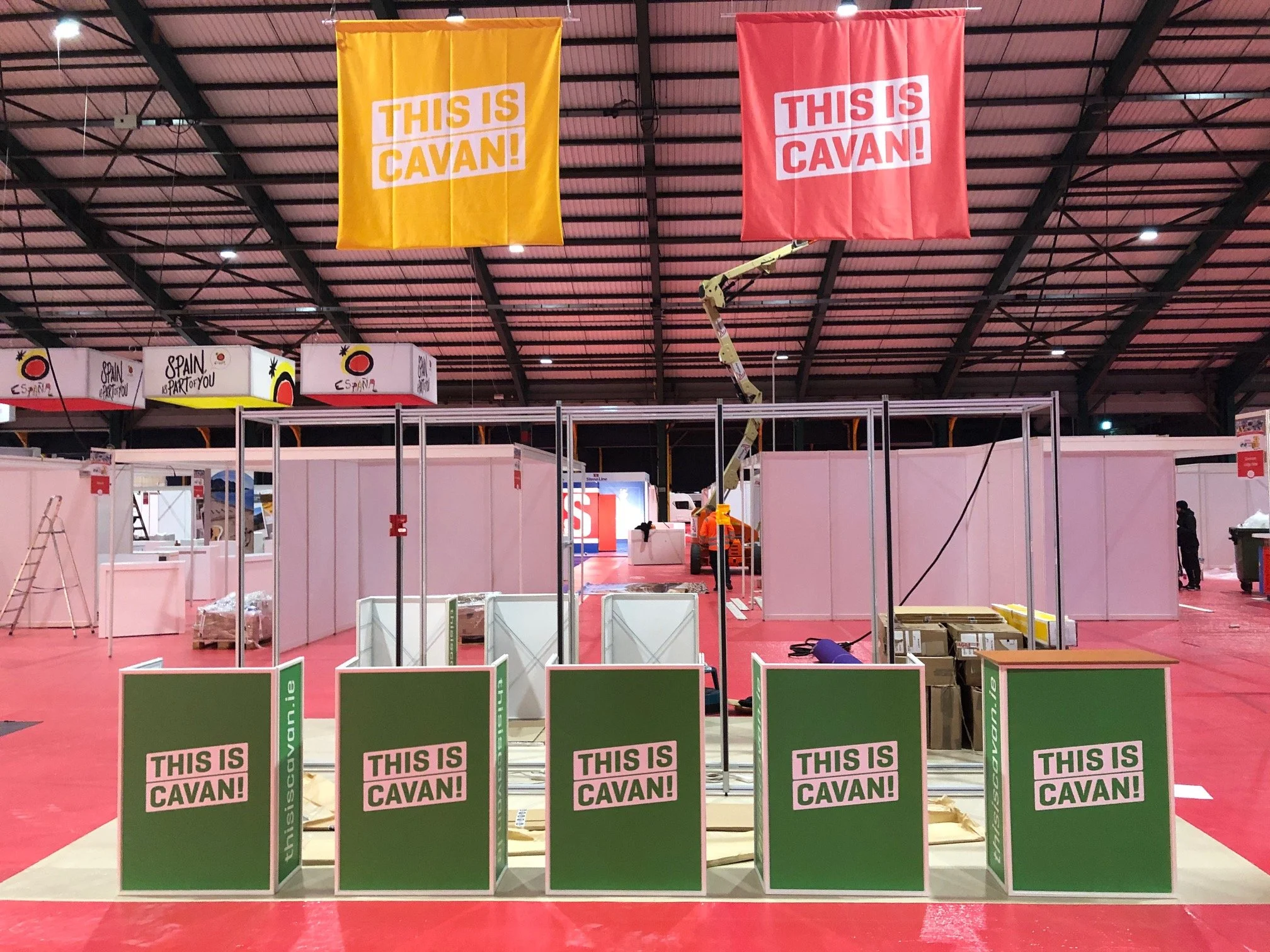 Indoor exhibition space with banners reading "This is Cavan!" hanging from the ceiling, partially constructed booths with green signs also reading "This is Cavan!", a ladder, a cherry picker, construction materials, and workers in safety vests.