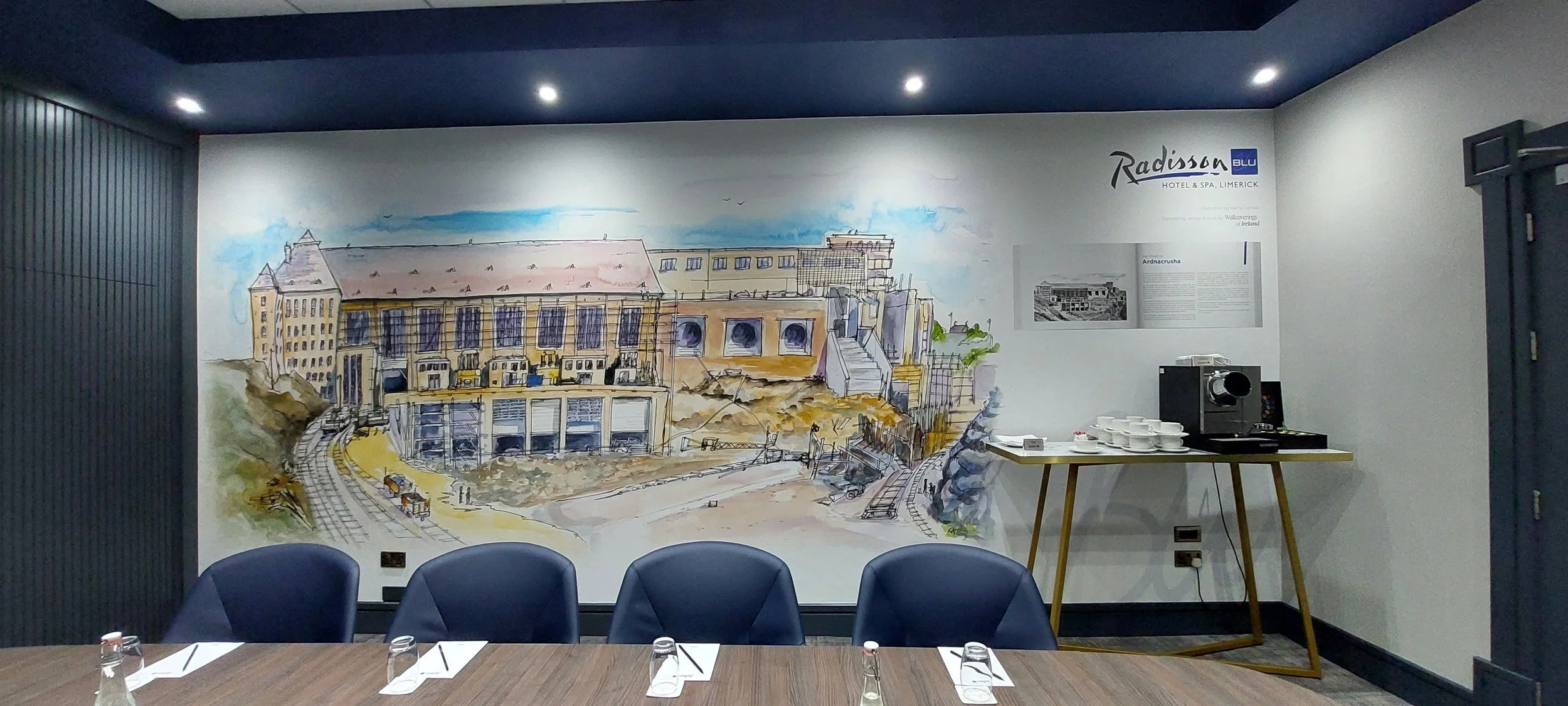 A painted mural of a historic building on a white wall in a conference room with blue chairs, a wooden table set with glasses and utensils, and a coffee station with cups and a coffee machine.