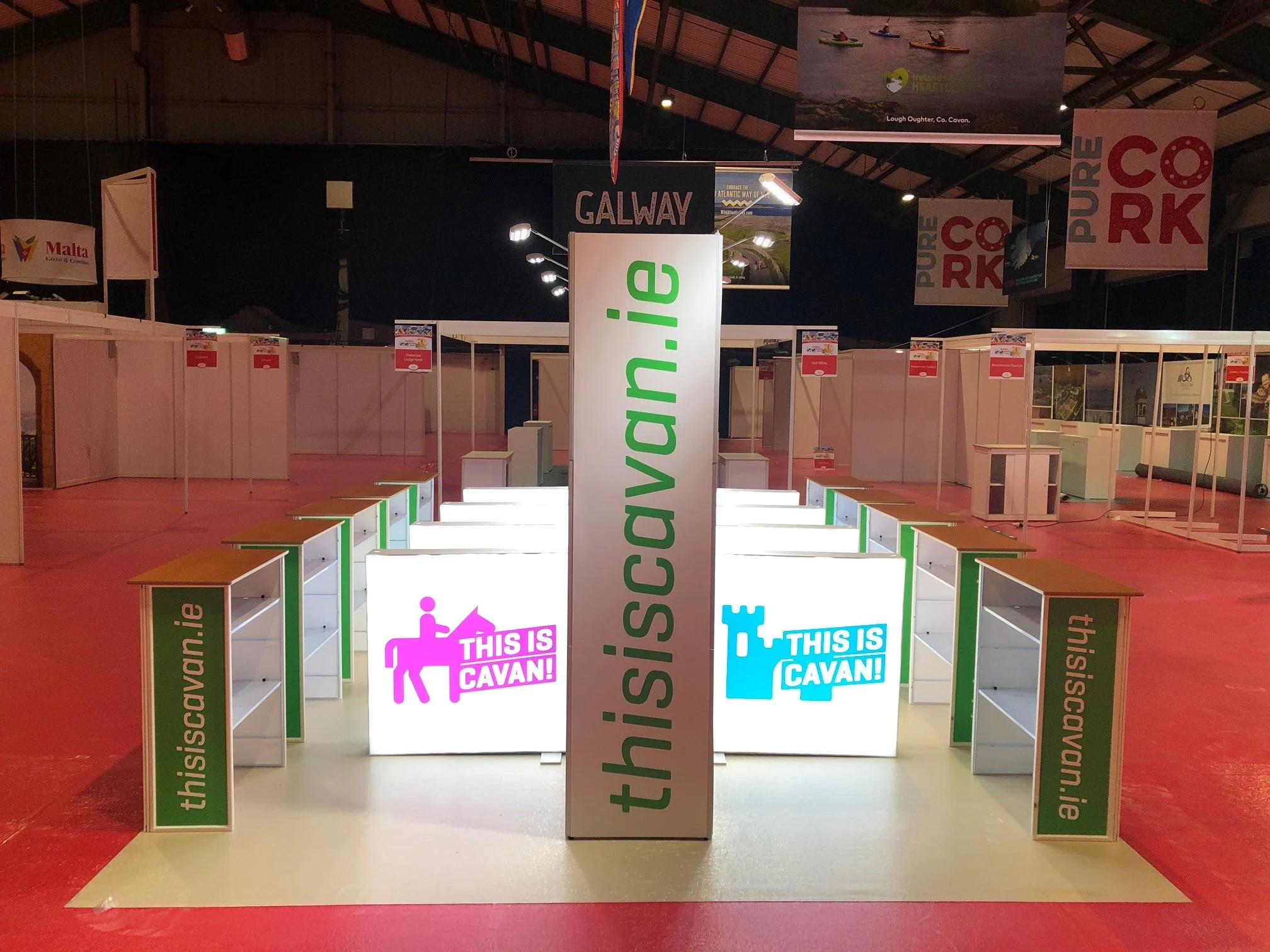 Booth at an exhibition with signs that read 'thisiscavan.ie' and 'THIS IS CAVAN!' with a pink illustration of a person on a horse and a blue representation of a castle, set in an indoor venue.
