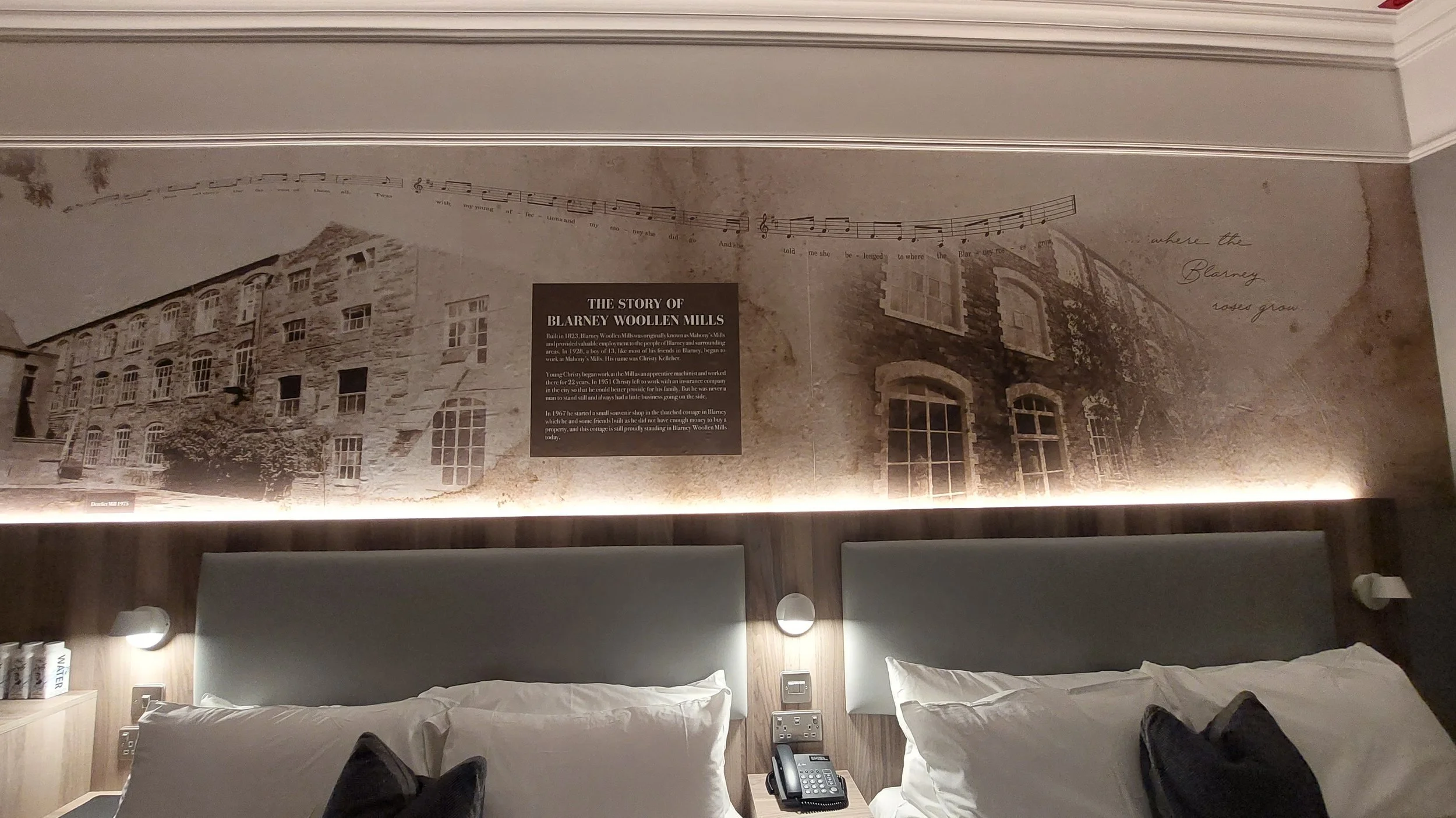 Hotel room with two beds, headboards, and a wall mural depicting the historic Blarney Woollen Mills building with an informational plaque and decorative text, dim lighting, and a telephone on the nightstand.
