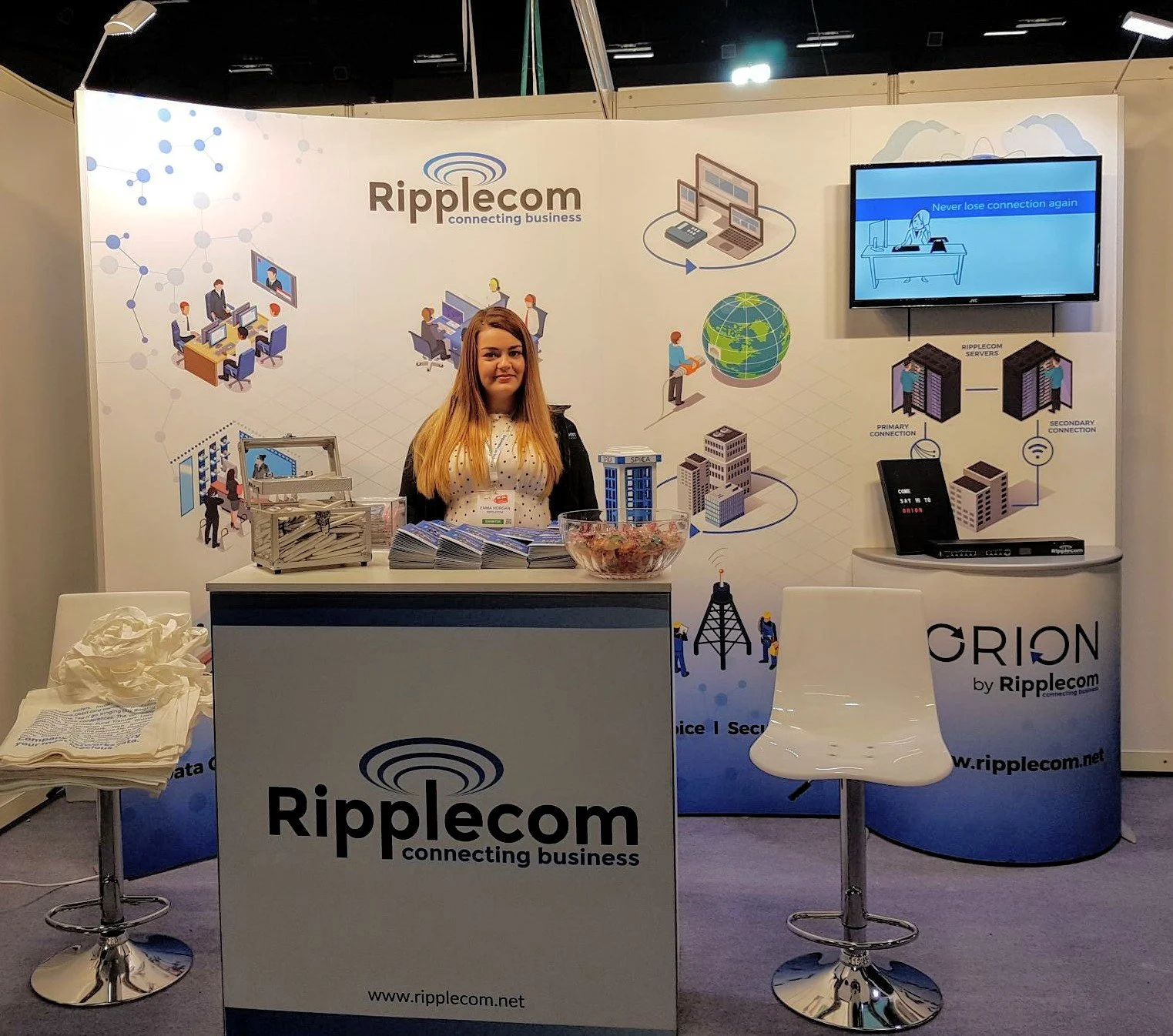 A woman standing behind a booth with Ripplecom branding at a trade show or conference, with promotional materials, a bowl of candies, and digital graphics on the backdrop showcasing internet connectivity and networking.