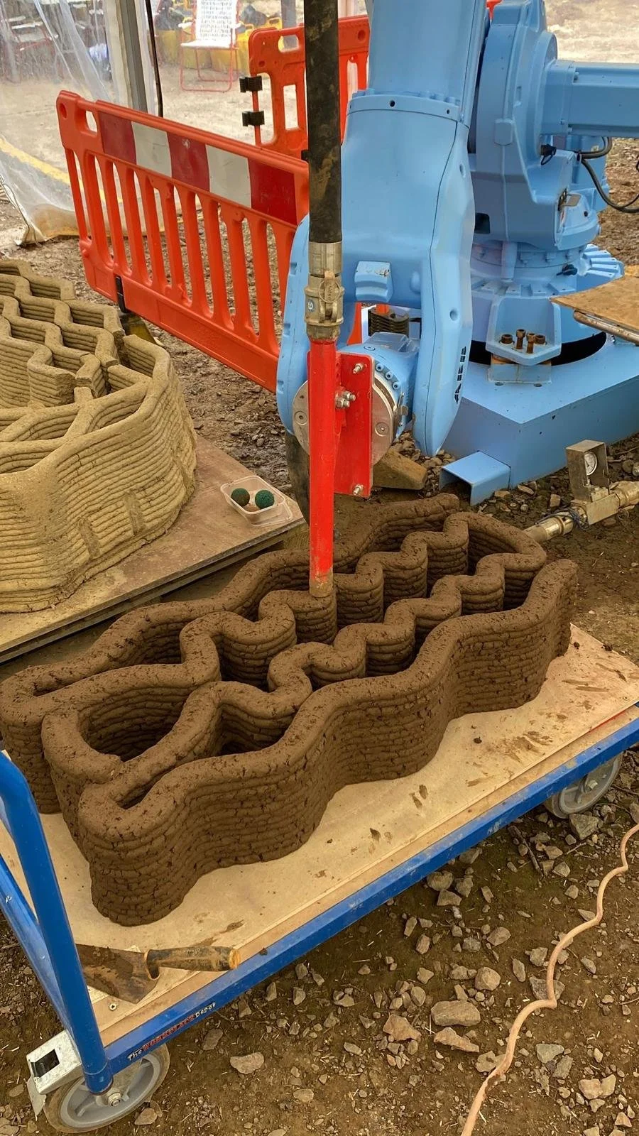 ABB robotic arm printing a sinusoidal double-skin earth wall section in progress at Earth Build Project's Stamford workshop. The layered toolpath geometry — forming interlocking cavities for thermal mass and structural performance — is visible mid-pr