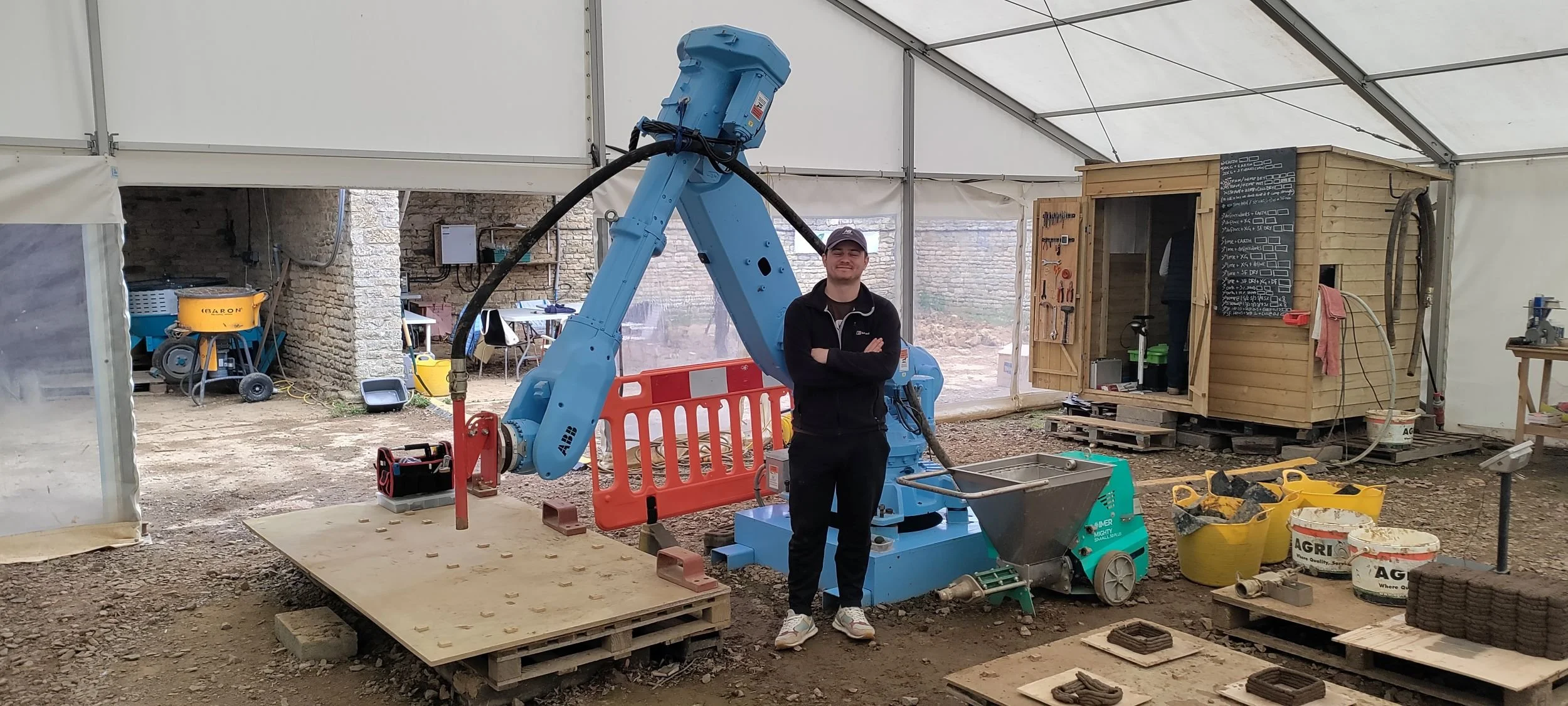 Earth Build Project's ABB robotic arm at the Stamford workshop, configured for 3DPE production. Print palettes, hopper, and material storage visible in the working fabrication environment. Image: Ben Scott.