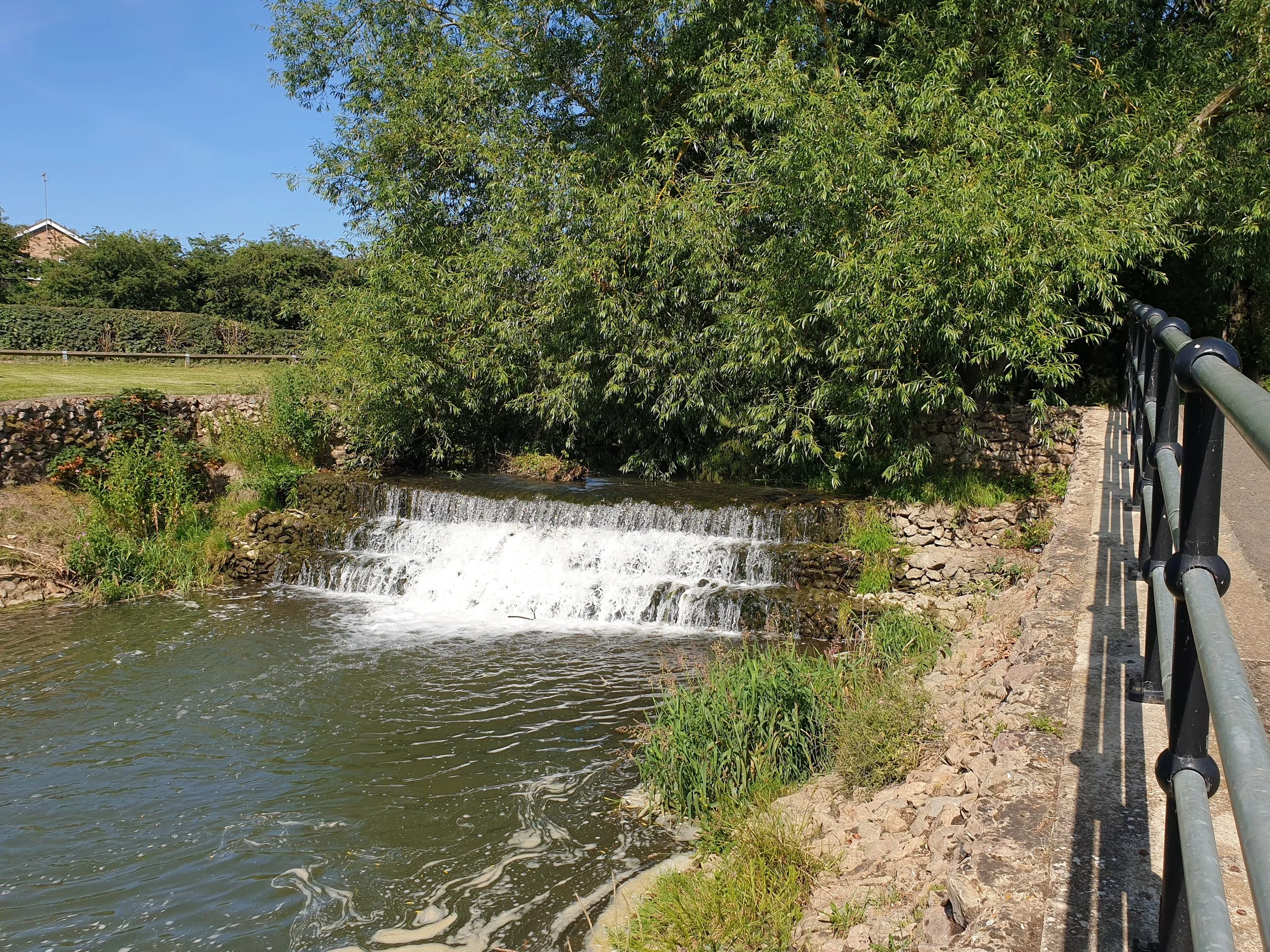 A small waterfall flowing over rocks in a creek, with a bridge and lush green trees in the background under a clear blue sky.