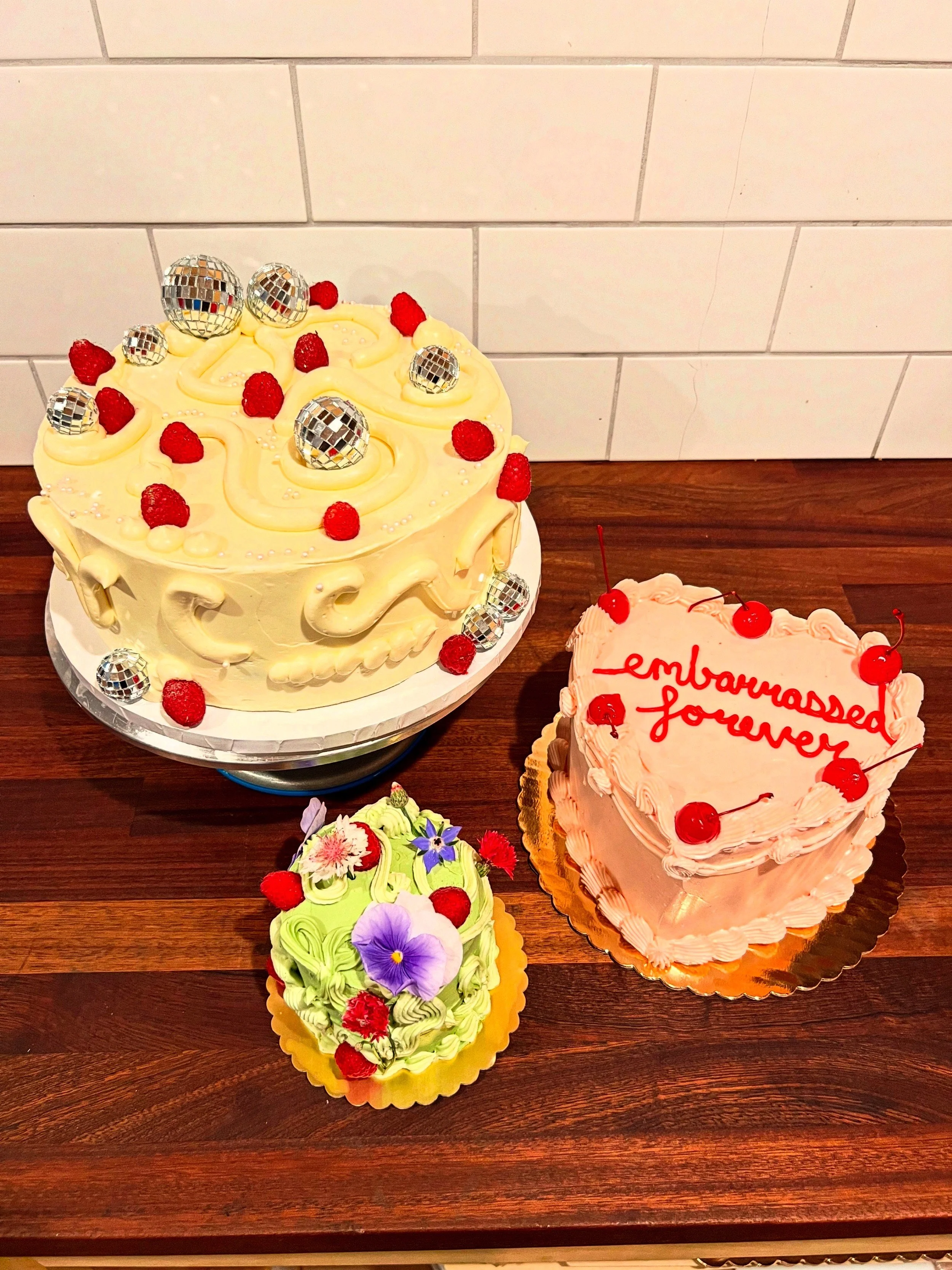 Trio of celebration cakes on a wood table, including a lemon buttercream cake with chocolate disco-ball spheres and raspberries, a pink heart-shaped cake, and a mini floral cake.