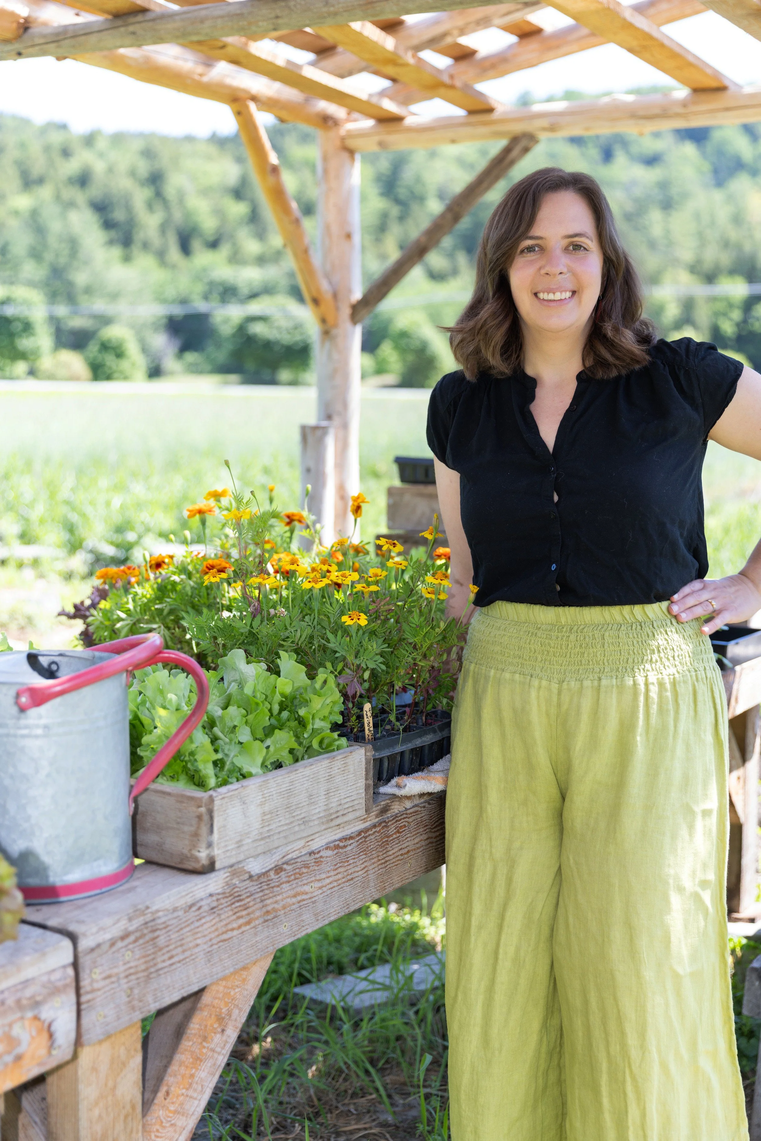 Sarah standing beside a wooden farm stand with marigolds, lettuce starts, and a watering can.