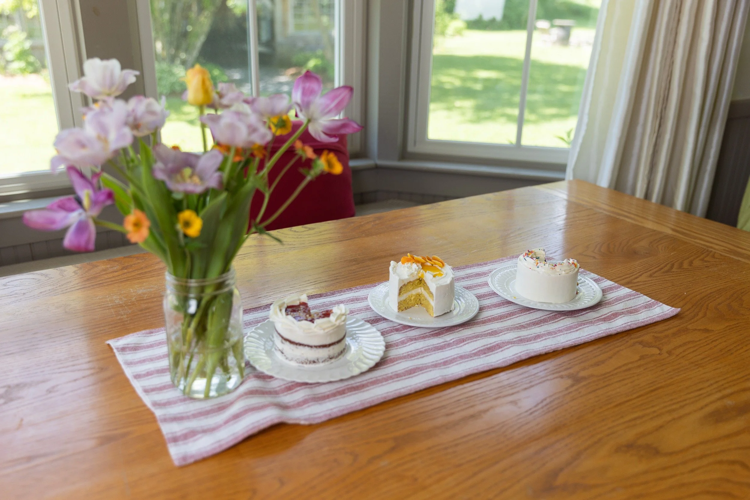 Three mini cake samples on a striped table runner with a vase of fresh tulips, set on a wood dining table.