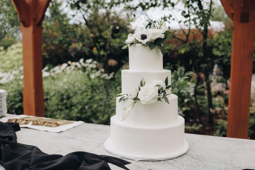 Four-tier white wedding cake with white anemones and greenery, displayed outdoors under a wood pergola. Goose Chase Cake Design | Weddings | Photo: Lillian Seiber Photography