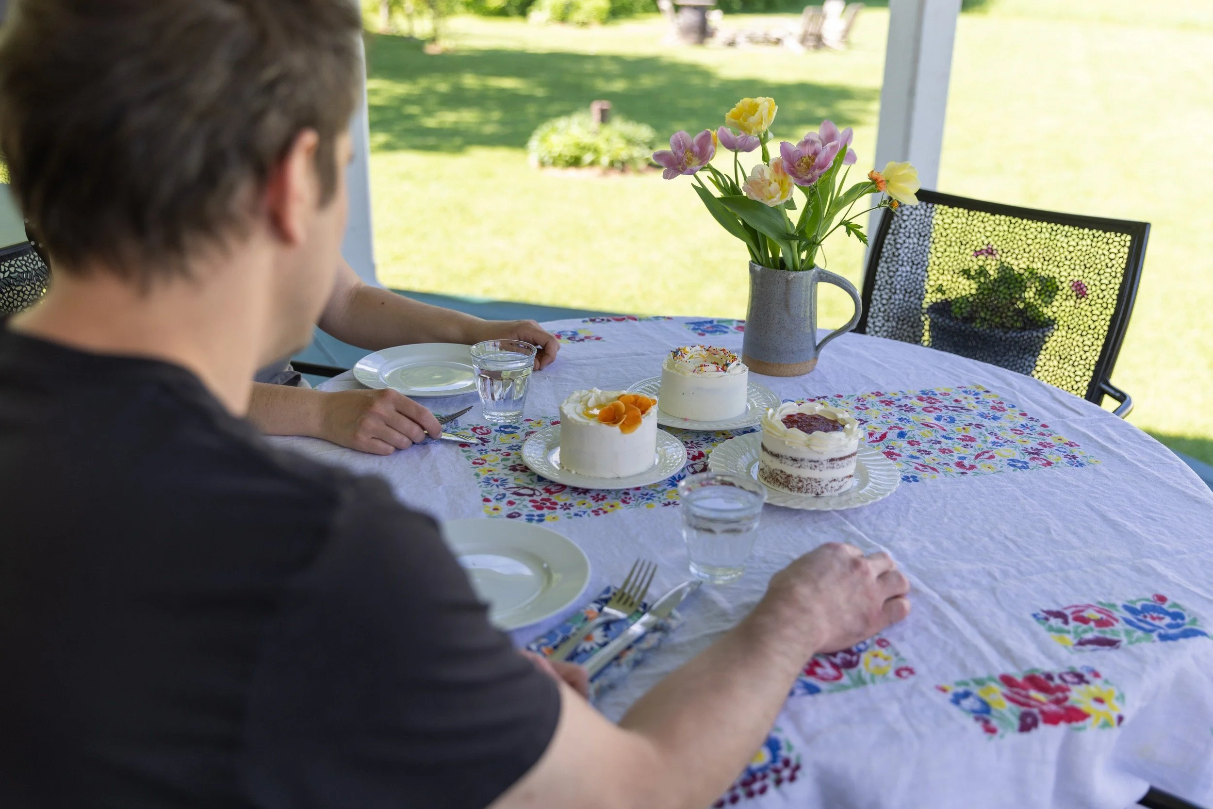 A guest reaching for a mini cake at an outdoor tasting table set with fresh tulips and a floral tablecloth.