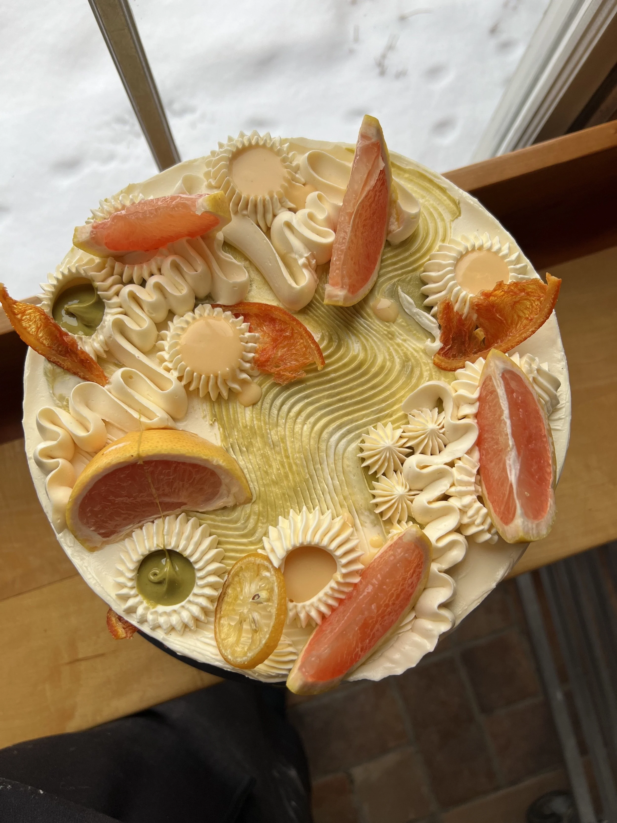 Overhead view of a celebration cake topped with citrus slices, dried orange wheels, and delicate piped rosettes in cream and gold tones.