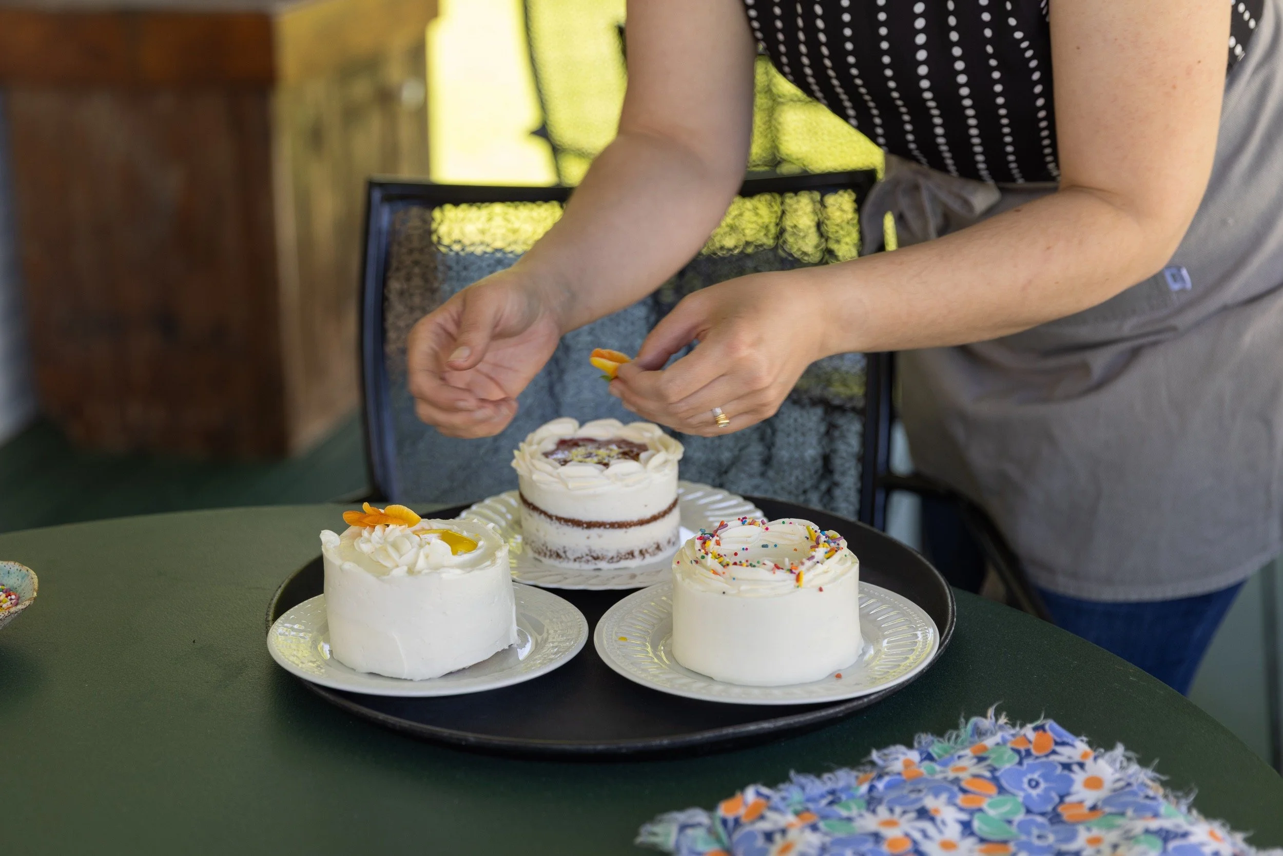 Close-up of hands decorating three mini celebration cakes with fresh flowers on an outdoor table.