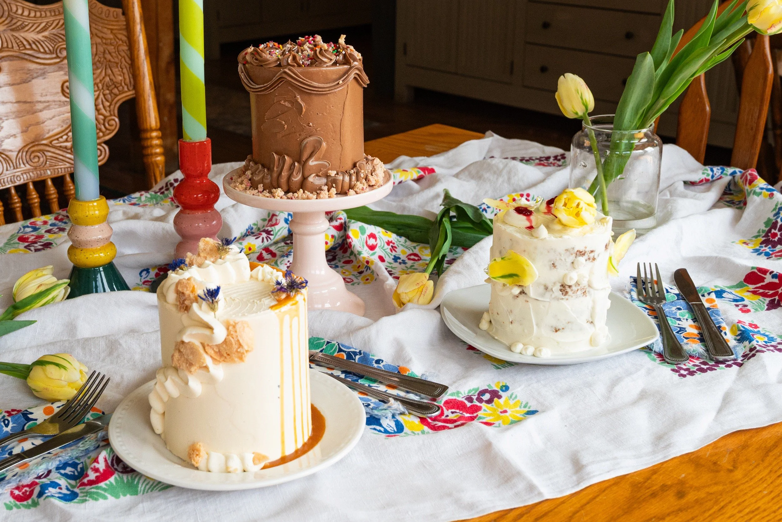 Trio of mini celebration cakes styled on a floral tablecloth with candles and tulips, including a chocolate cake, a caramel cake, and a floral-topped cake.