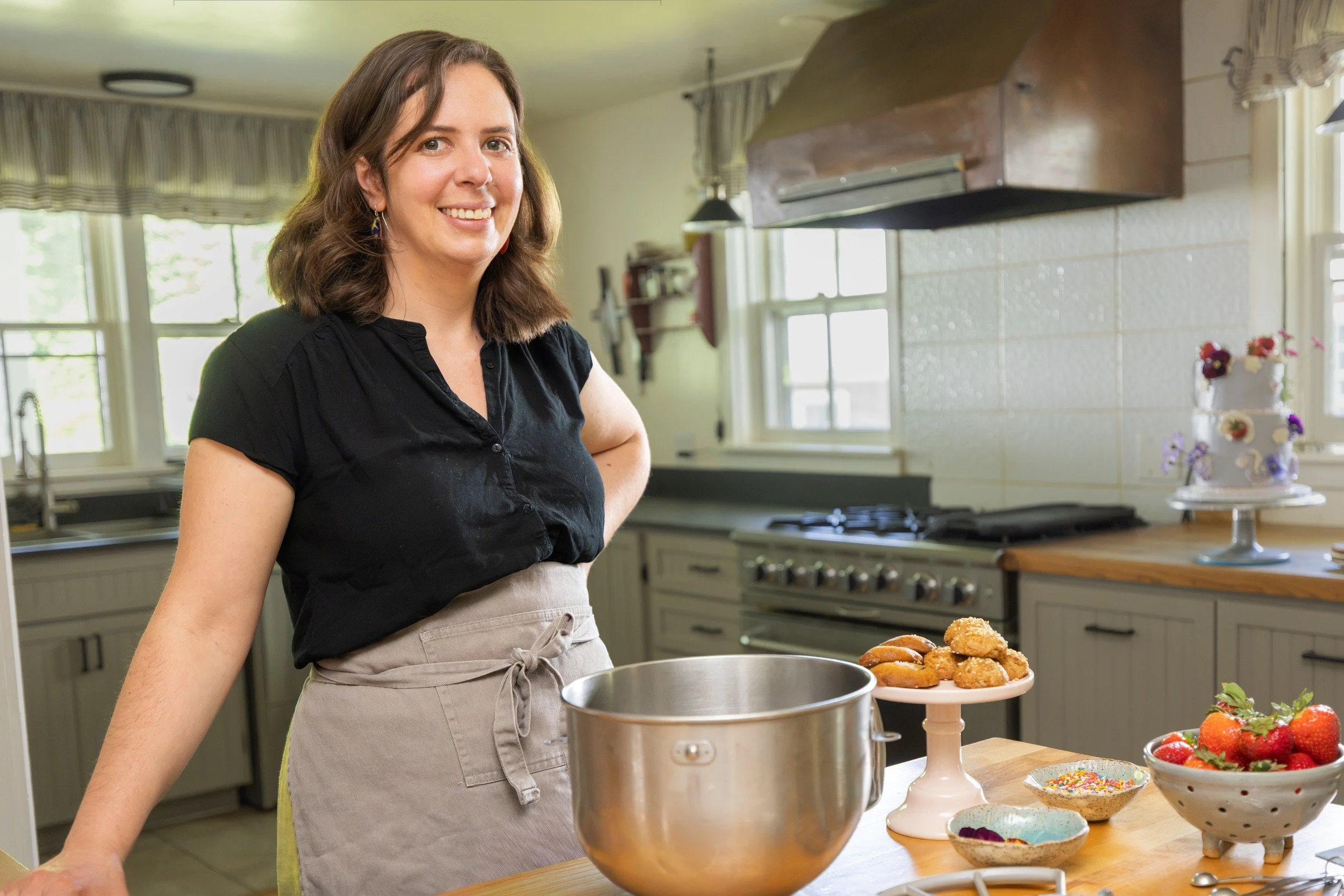 Sarah smiling in her kitchen beside a stand mixer, cookies, and a bowl of fresh strawberries.