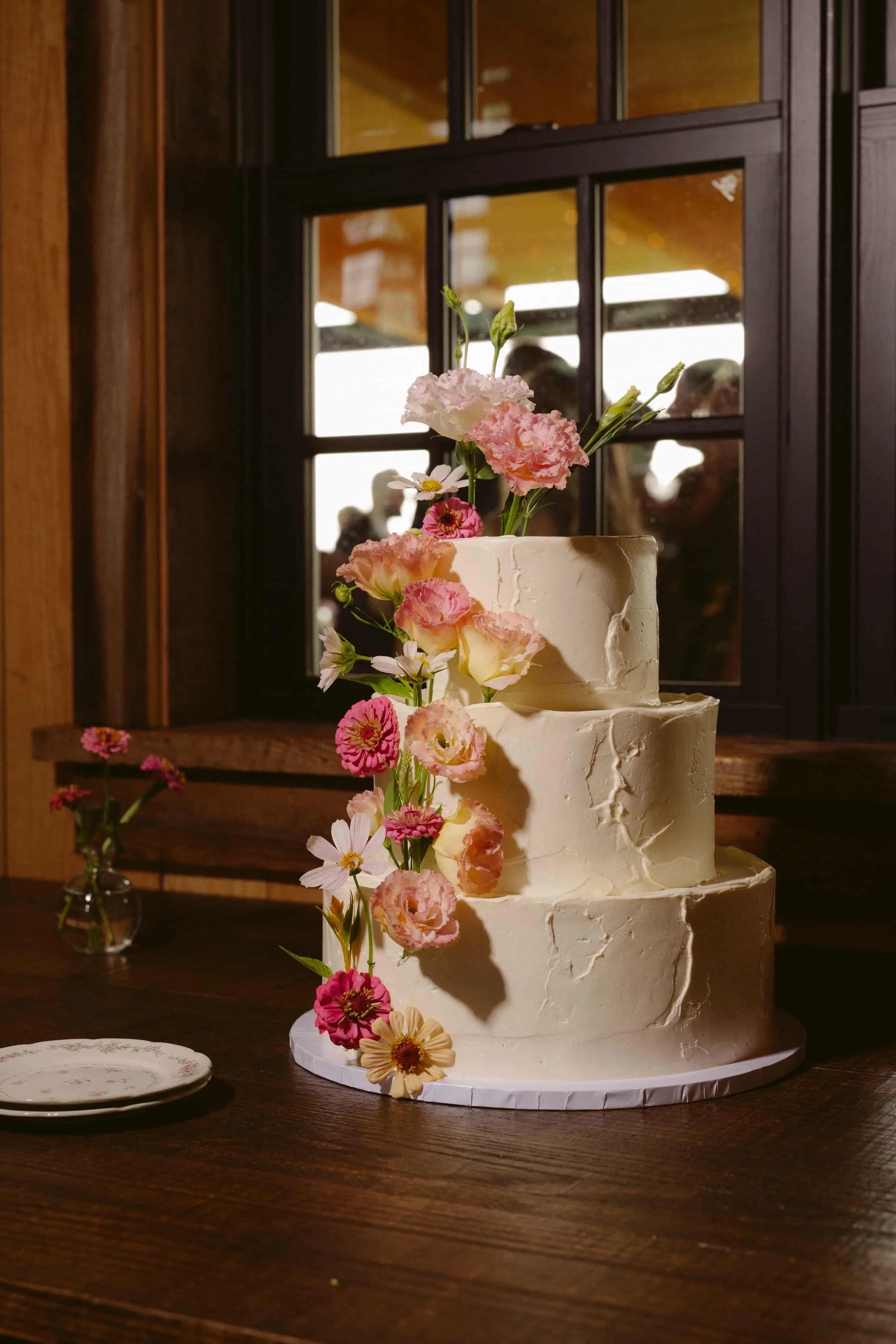 Three-tier textured wedding cake with cascading pink and coral florals, displayed near a window. Goose Chase Cake Design | Weddings | Photo: Patrick McCormack