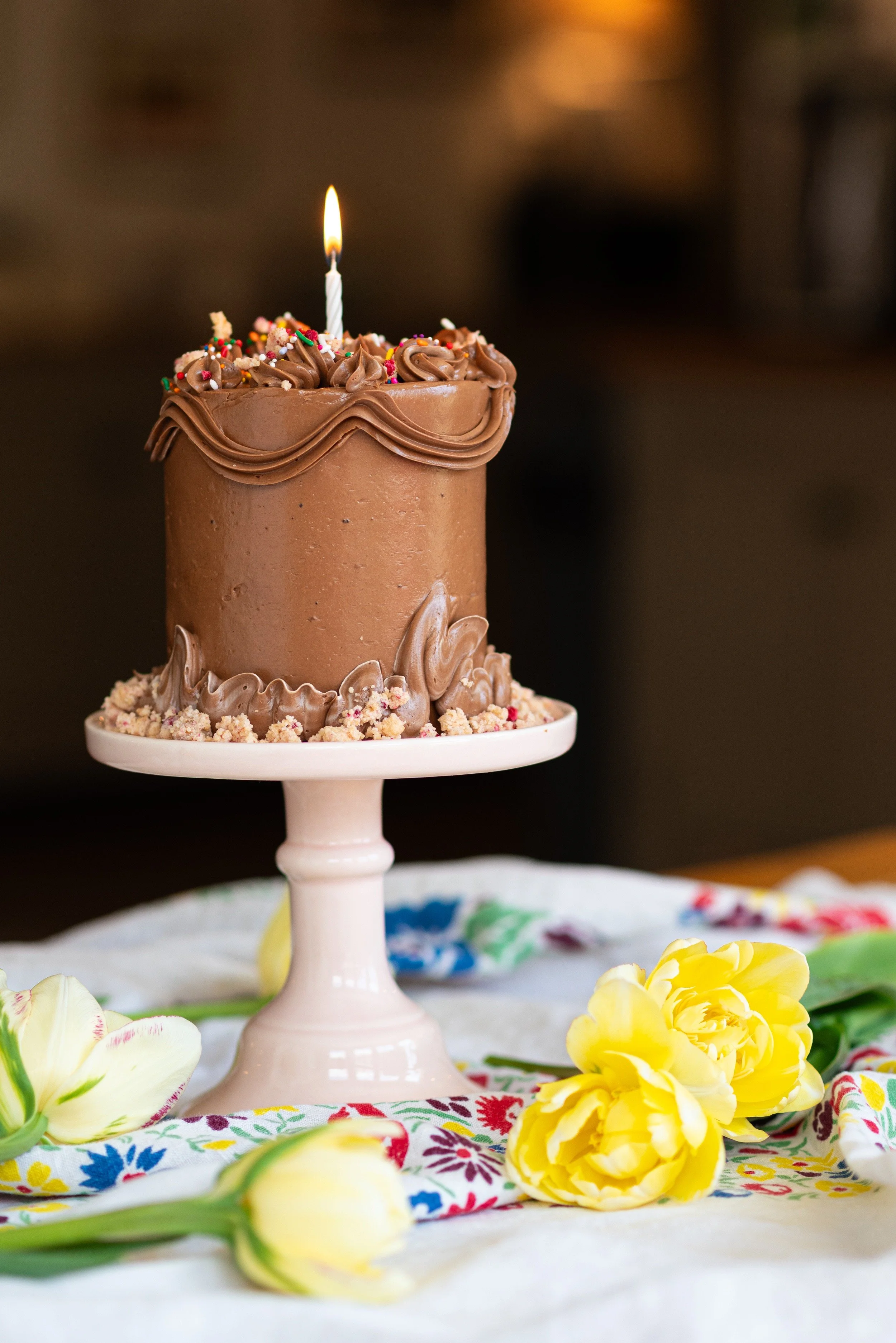 Mini chocolate celebration cake with a lit birthday candle, crumb topping, and fresh yellow tulips, displayed on a pink cake stand.