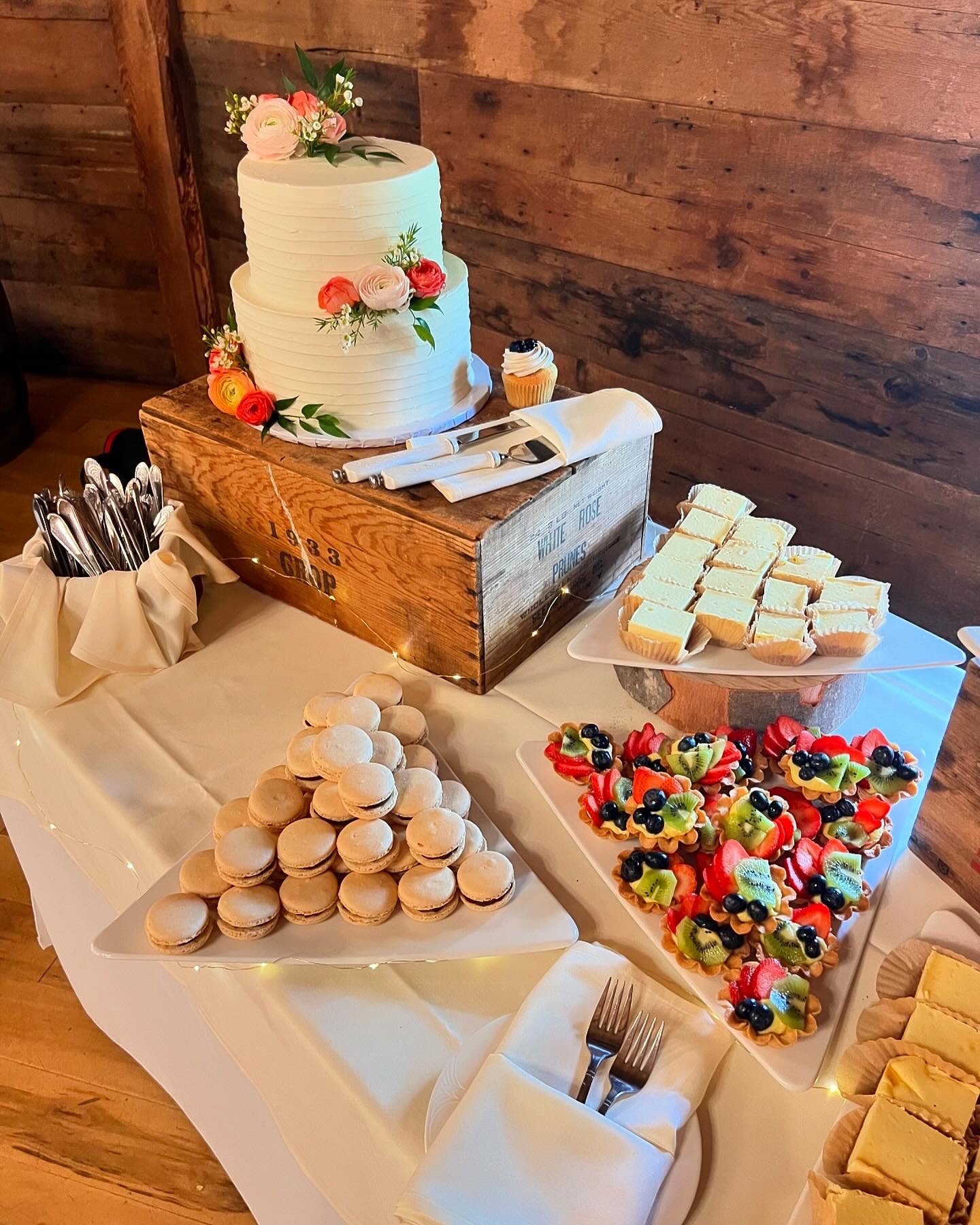 Two-tier white buttercream wedding cake with fresh flowers on a rustic wood cake stand, displayed alongside mini cupcakes, macarons, and fruit tarts at a Vermont wedding dessert table.
