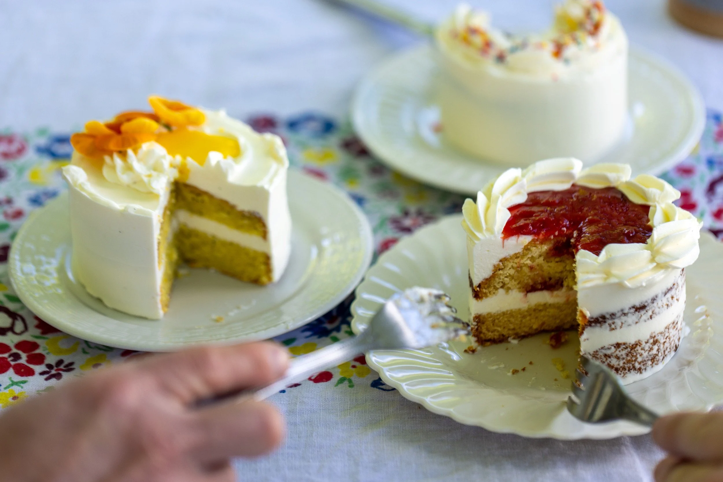 Close-up of two mini cake samples, one citrus-topped and one filled with berry jam, sliced open on a floral tablecloth.
