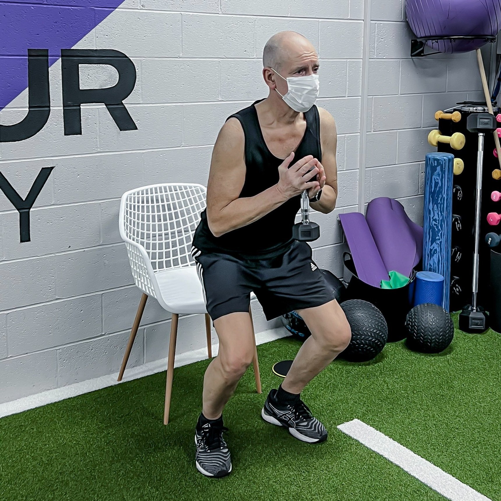 An older woman in workout attire, wearing a face mask, sitting on a white chair, holding a dumbbell in a squat position in a gym.