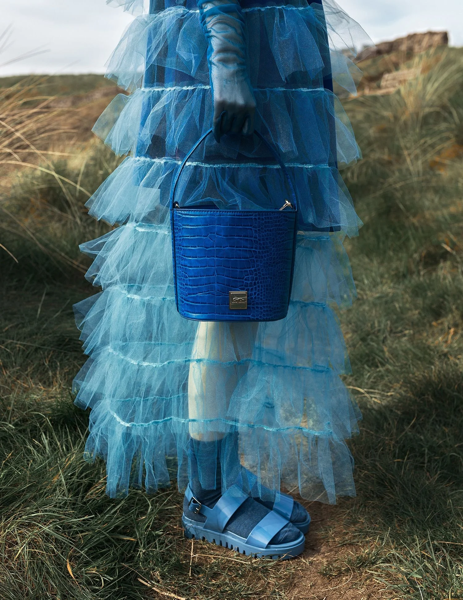 Person wearing a blue layered tulle gown, black gloves, and sporting blue sandals, holding a blue crocodile-embossed handbag, standing outdoors on grassy terrain.