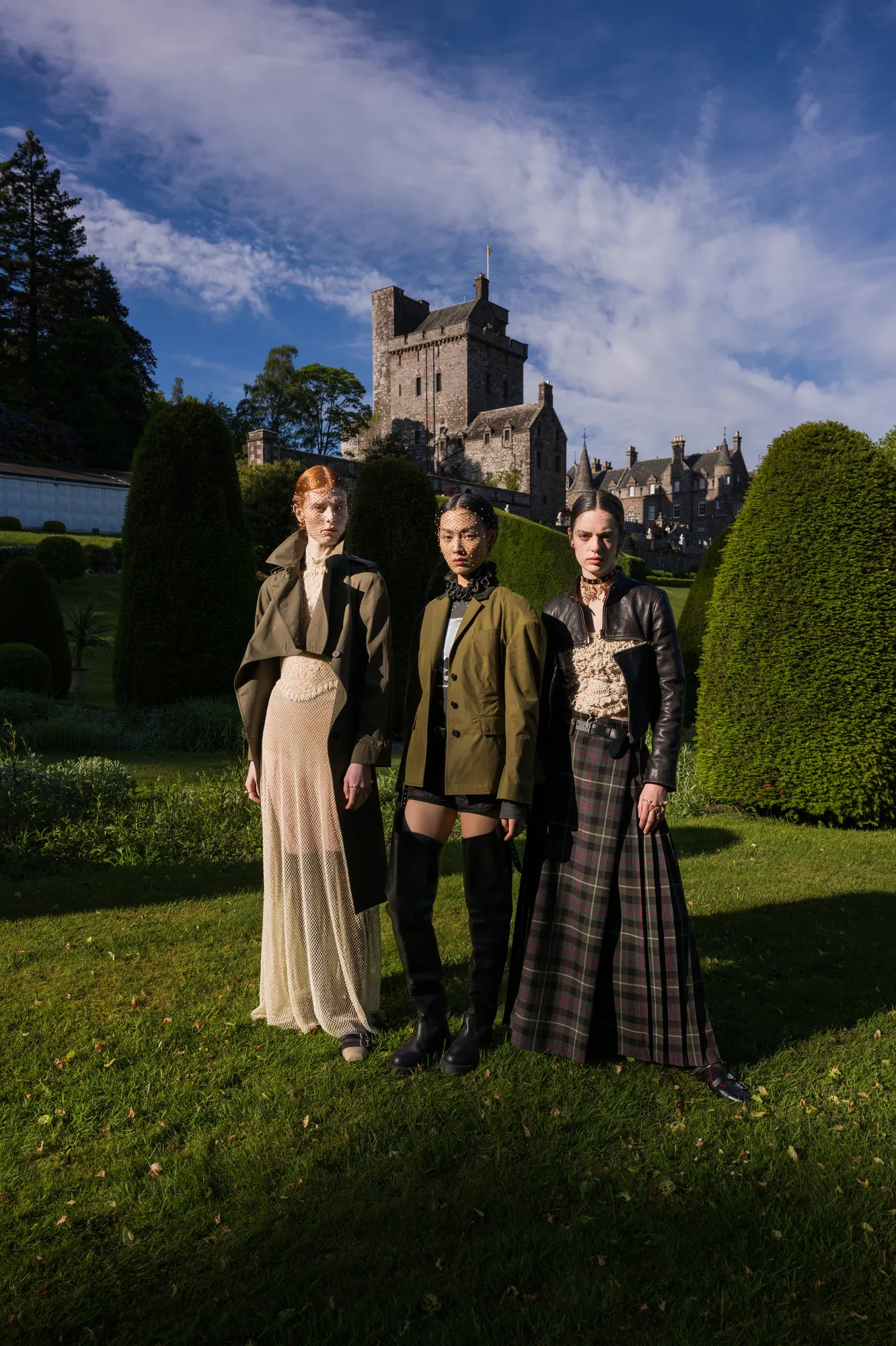 Three women standing outdoors on a lawn with a castle in the background. They are dressed in fashionable, eclectic outfits and posing confidently.