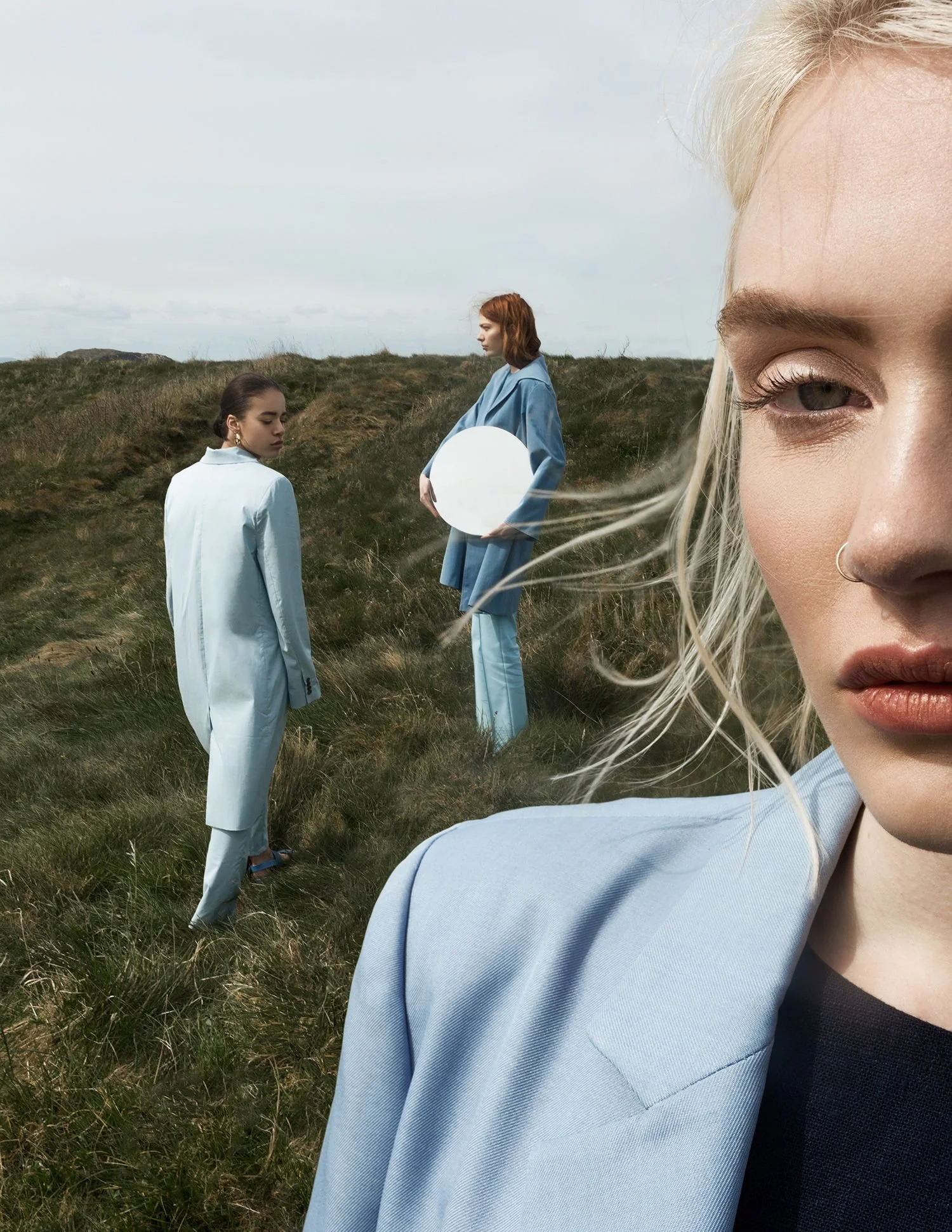 Close-up of a woman with blonde hair, wearing a light blue blazer, standing outdoors in a grassy area with two other women in blue outfits and one holding a mirror in the background.