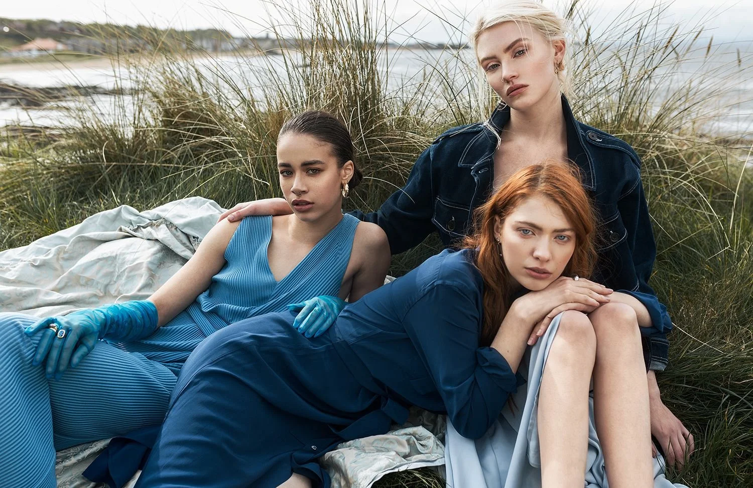 Three women sitting outdoors on a blanket among tall grass with a body of water in the background, wearing stylish blue and denim outfits.