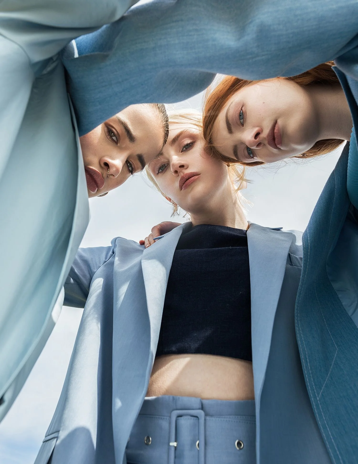 Three women with colorful hair and fashion outfits looking down at the camera from beneath, outdoors.