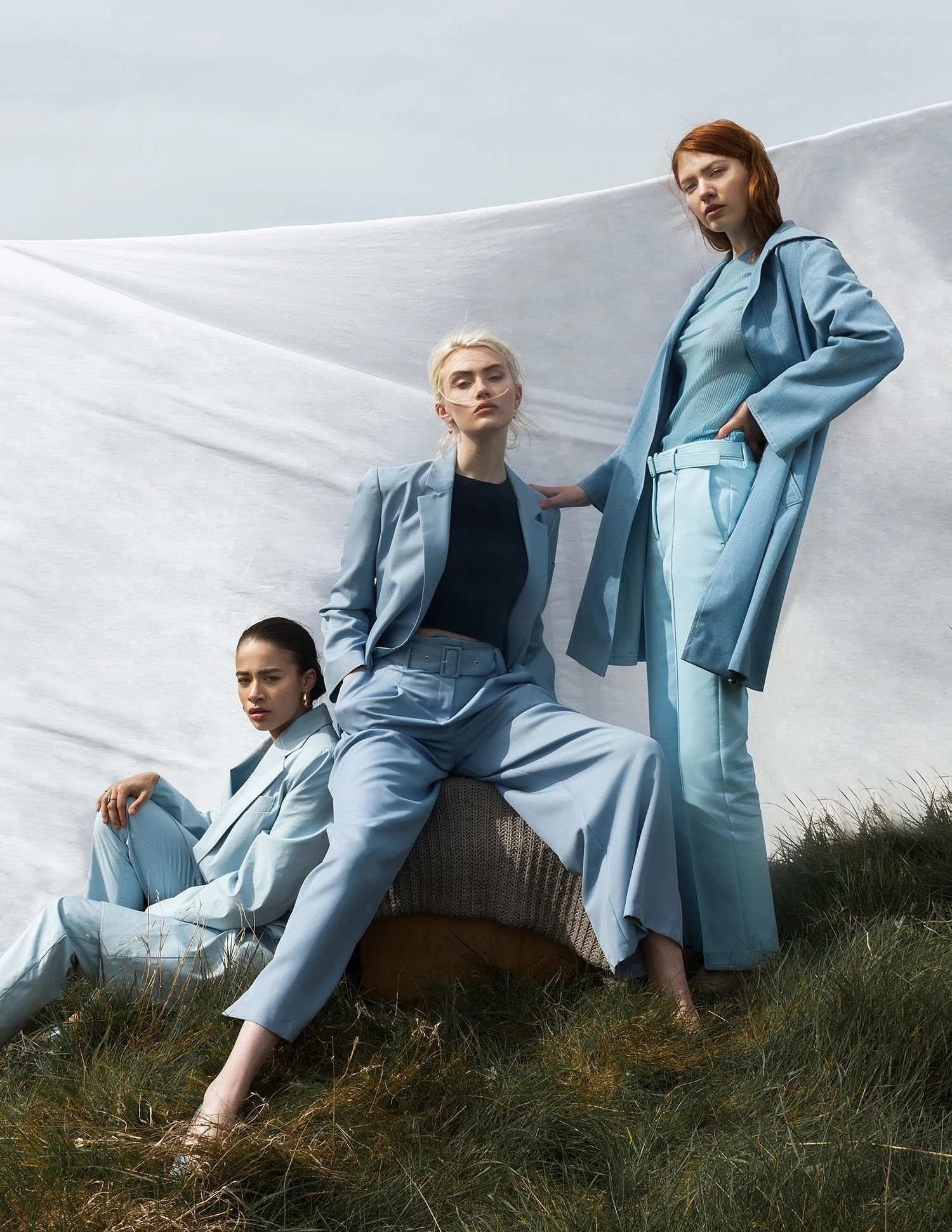 Three women dressed in light blue suits and casual clothing posing outdoors on grass against a white fabric backdrop.