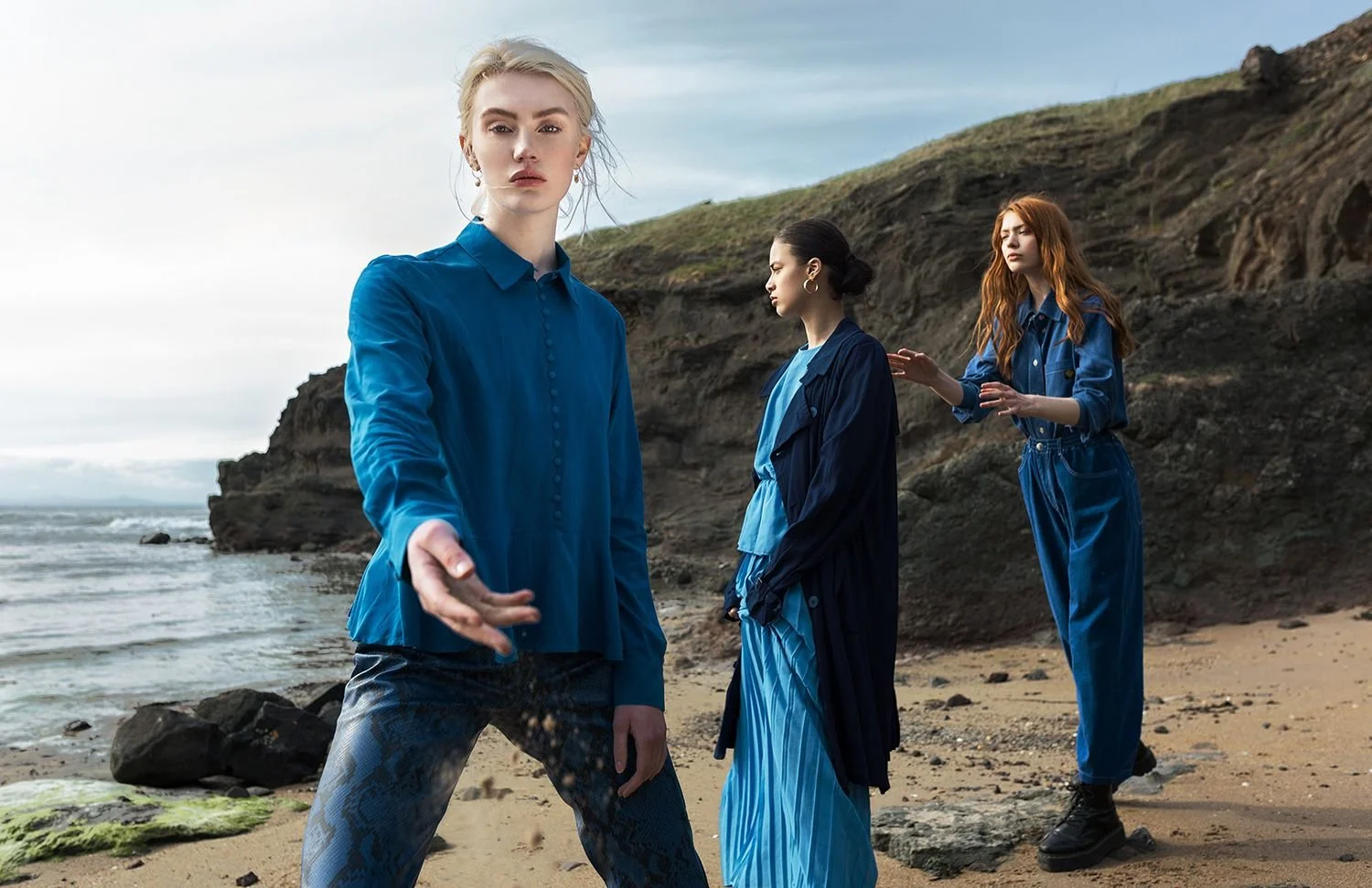 Three women in blue clothing stand on a beach with rocky cliffs in the background, one woman in the foreground reaching toward the camera.