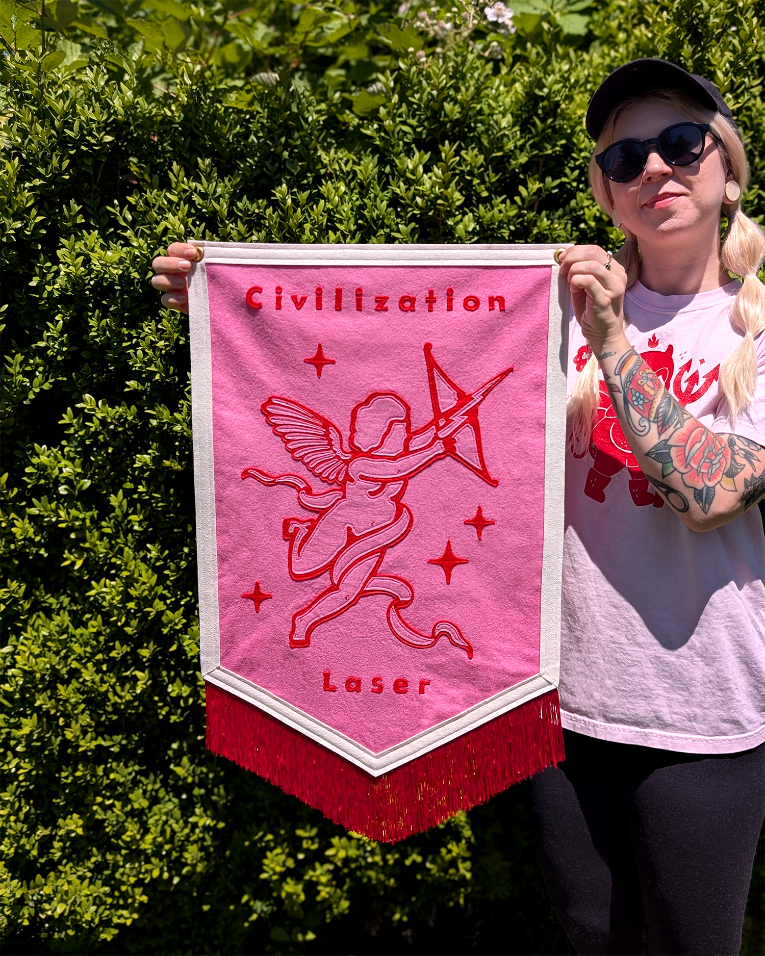 A woman holding a pink and red felt banner with a design of a cupid with a bow and arrow, surrounded by stars. The banner has the words 'civilization' at the top and 'Laser' at the bottom. The woman is wearing sunglasses, a gray cap, and a white t-sh