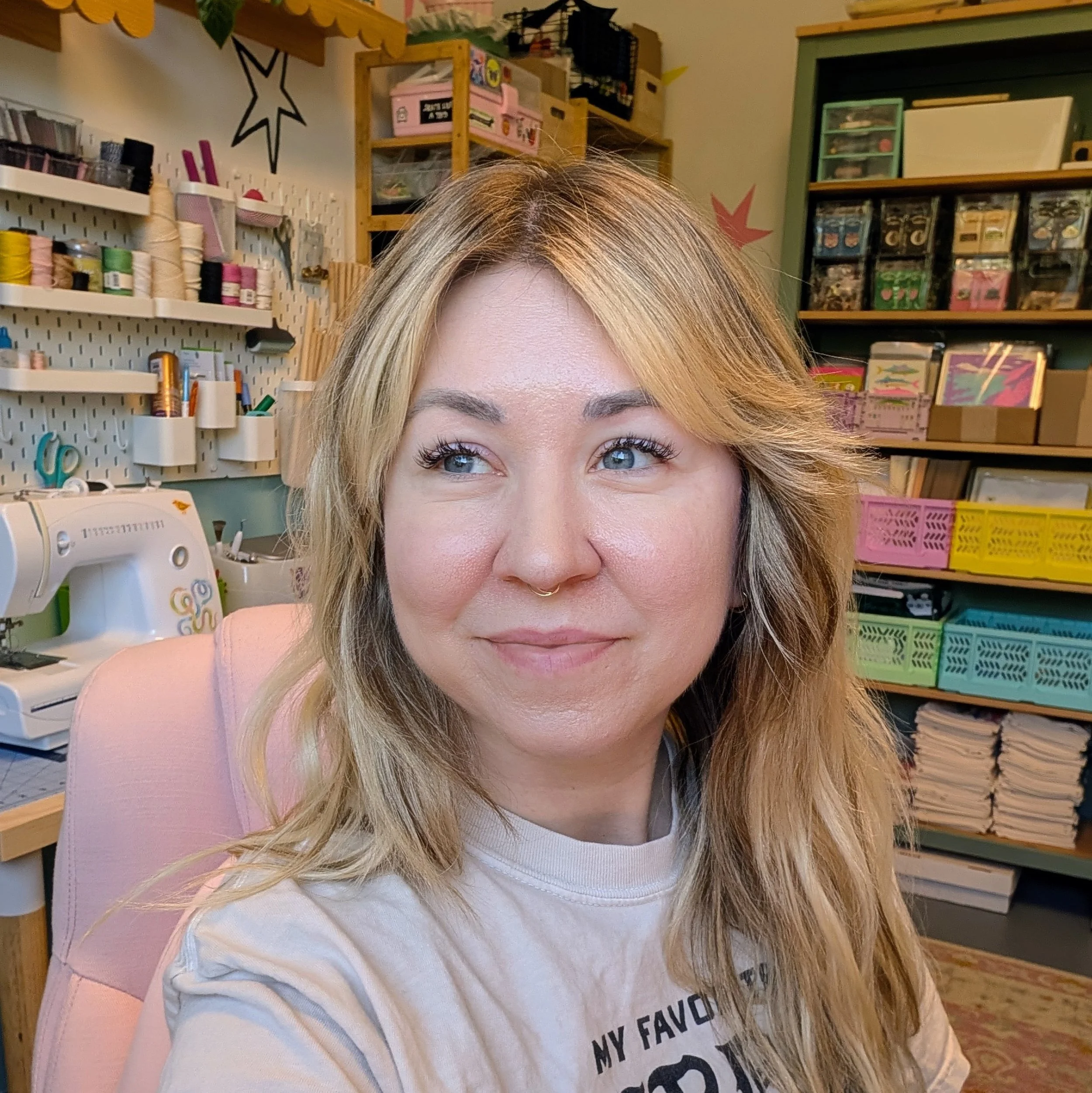 Close-up of a woman's face with long, blonde hair and blue eyes, in a craft studio space room with shelves of colorful supplies and organized boxes in the background.