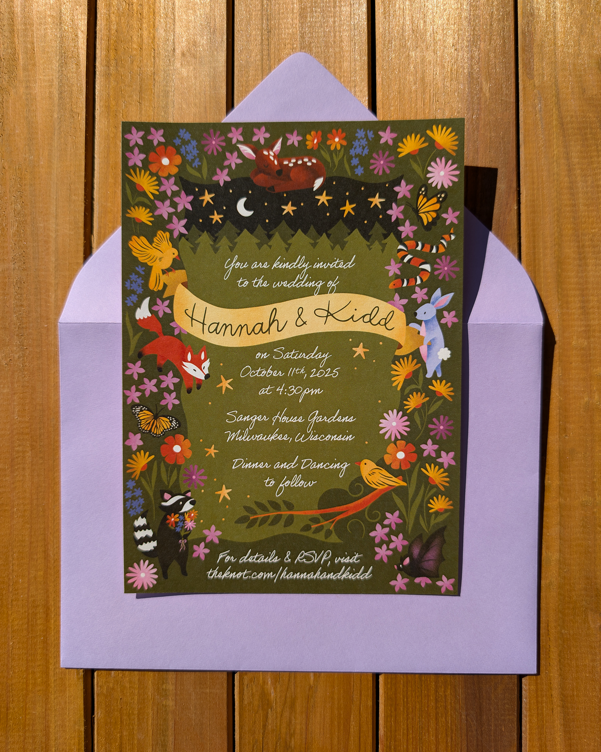 A forest-themed wedding invitation with colorful hand drawn illustrations of forest animals, flowers, and stars on a black background, set on a wooden surface with lavender envelope behind it.