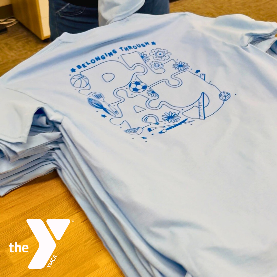 A stack of light blue T-shirts with a screenprinted graphic and the words 'Belonging Through PLAY', featuring illustrations of sports equipment, flowers, and toys. The design was made for the YMCA Columbia-Willamette kid's summer camp t-shirt design.