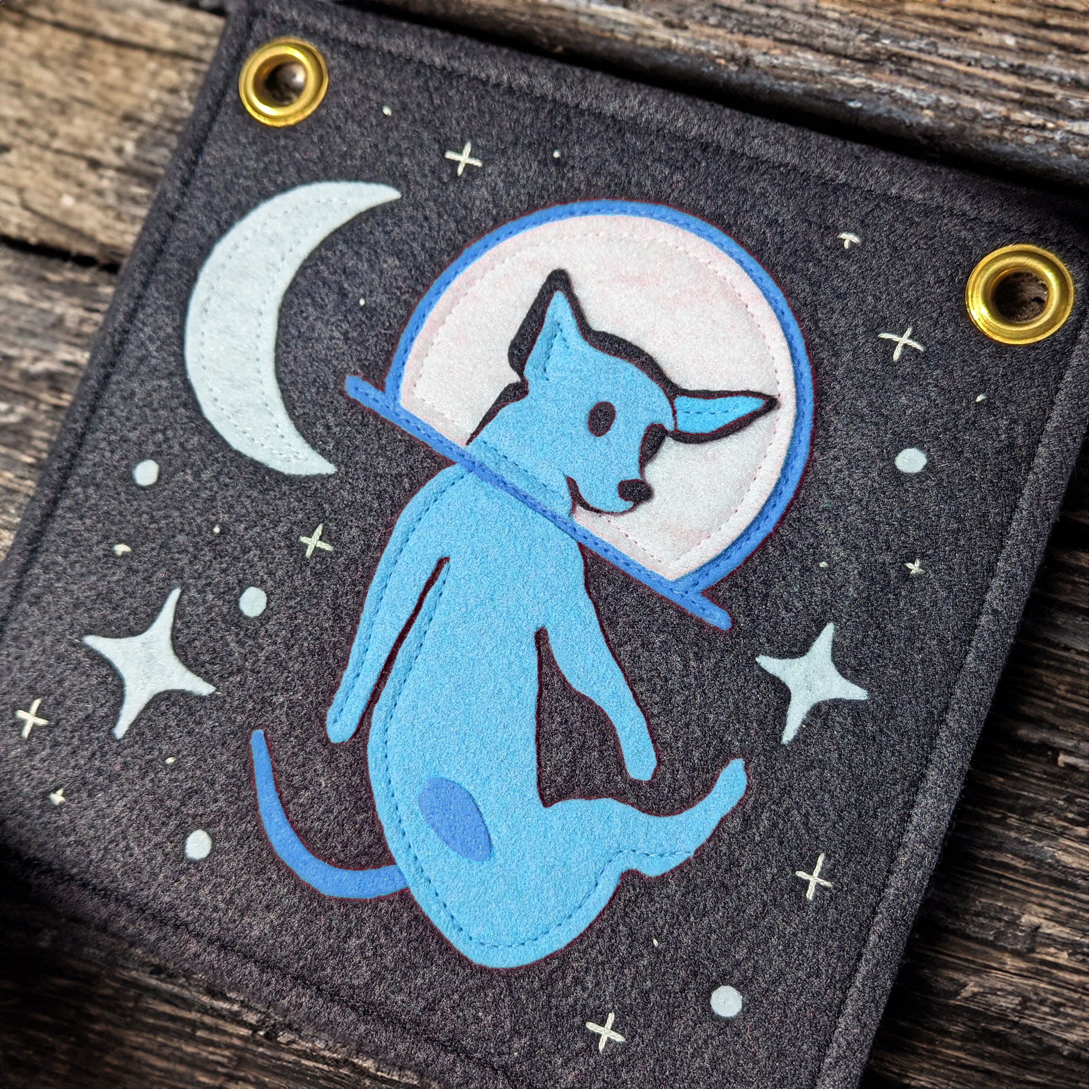 Close-up of a patch featuring a blue dog with a black nose inside a white circle, set against a black background with small white stars and a crescent moon. The patch is attached to a dark, textured fabric with gold eyelets.