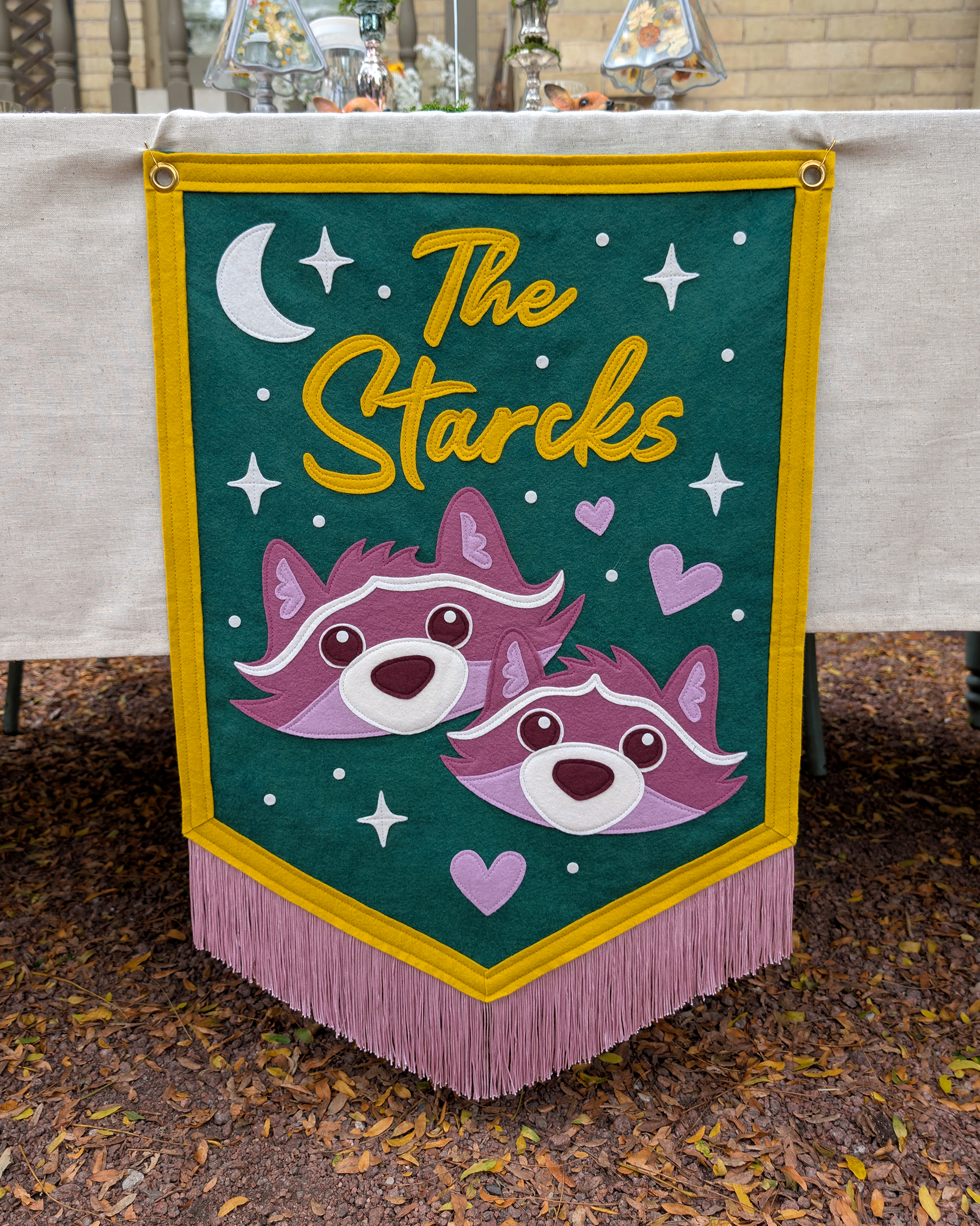 A colorful felt handmade wedding banner with cartoon raccoon faces, hearts, and white stars against a green background, surrounded by a yellow border and hanging from a table with lavender fringe at the bottom. Text reads "The Starcks" at the top.