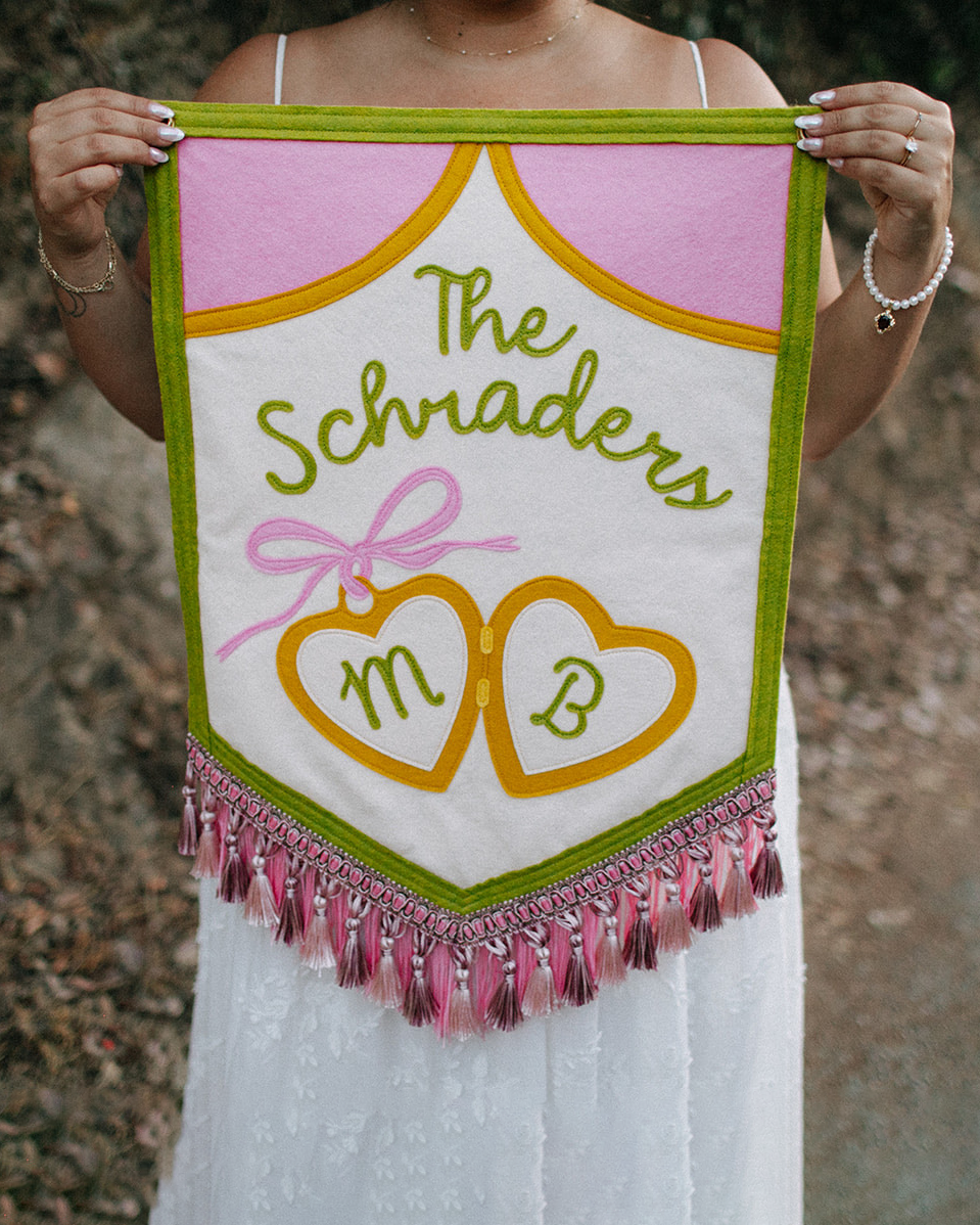 A woman holding a pink and white embroidered banner with green and yellow accents, decorated with pink tassels. The banner reads "The schaders" with two intertwined hearts and initials "M" and "B" inside each heart.
