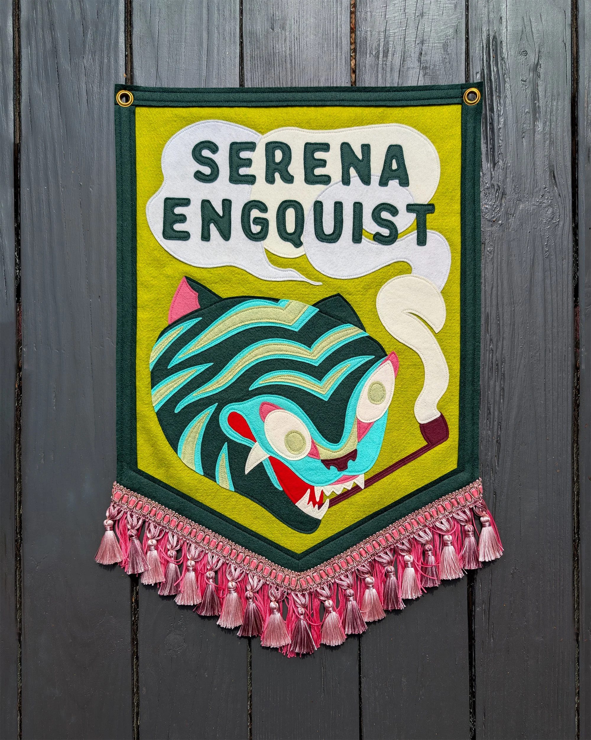 Colorful handmade textile hanging banner with pink tassel fringe, featuring the a smoking tiger tattoo design, made in colorful felt and the words 'Serena Enquist' in speech bubbles.