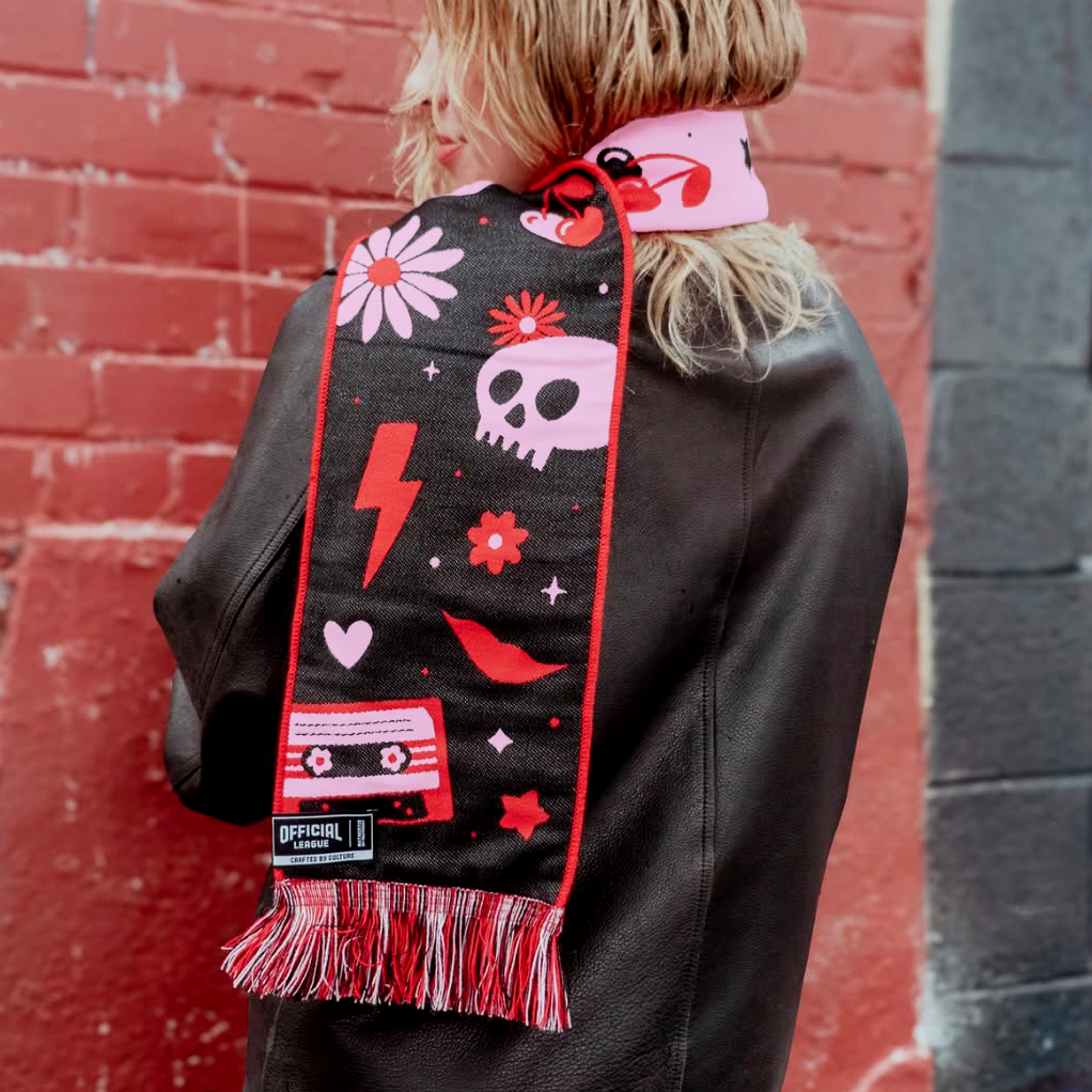 A person with blonde hair shown from the back wearing a black jacket and a pink and red soccer scarf with various handdrawn designs including a skull, flowers, a lightning bolt, a cassette tape, and a lip print, standing in front of a red brick wall.